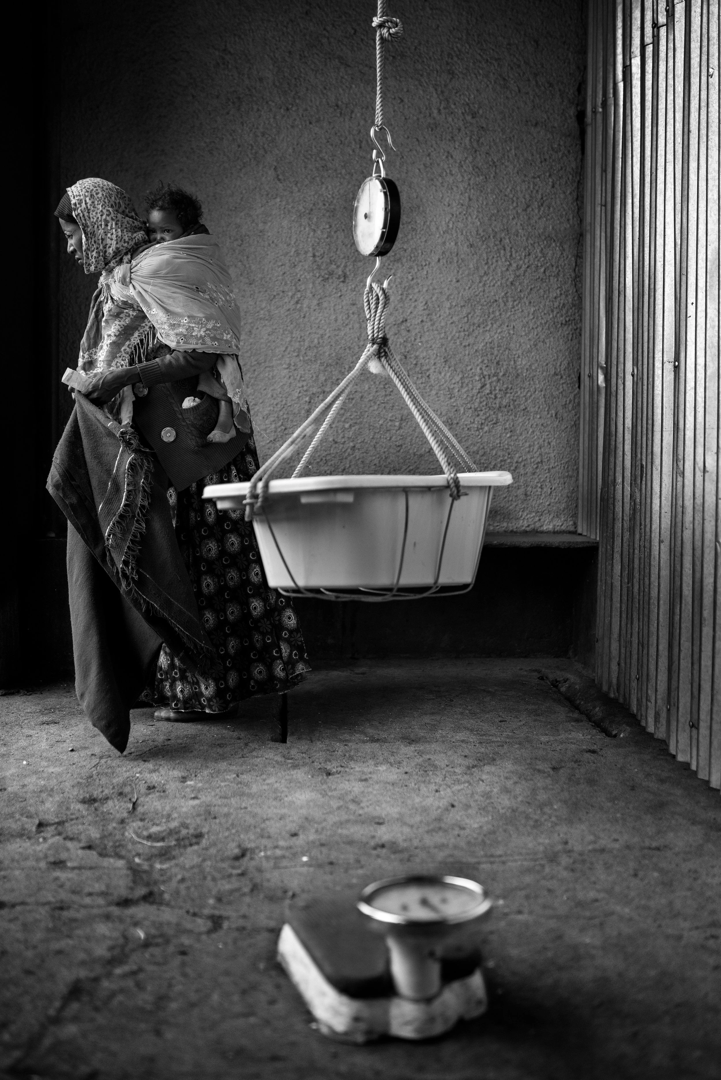 Mujer caminando mientras lleva a un niño en la espalda, con una balanza sobre el suelo y una bañera colgando en una cuerda en una pared de fondo.