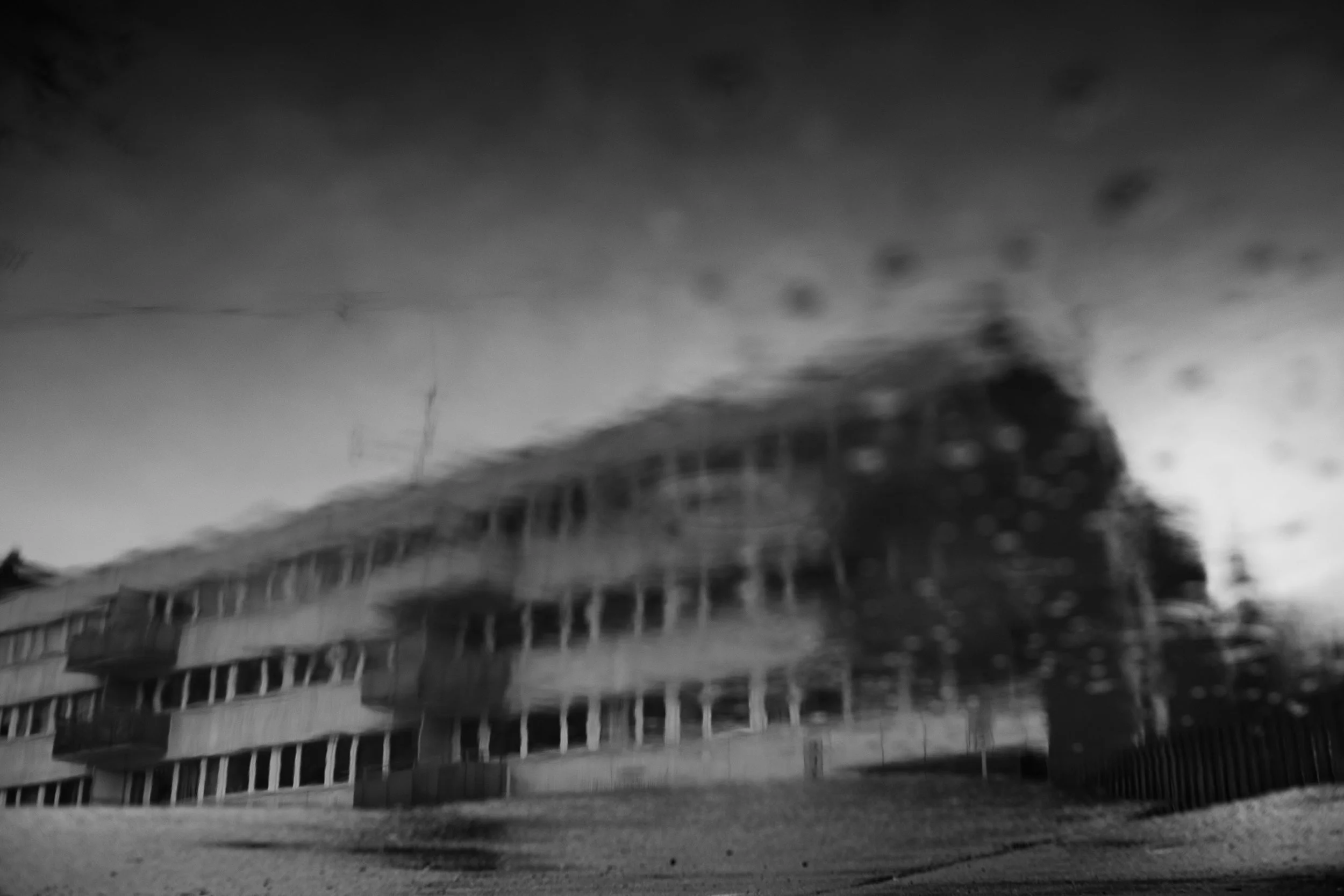 Edificio de varios pisos con ventanas y balcones, reflejado en un charco de agua en un día nublado.