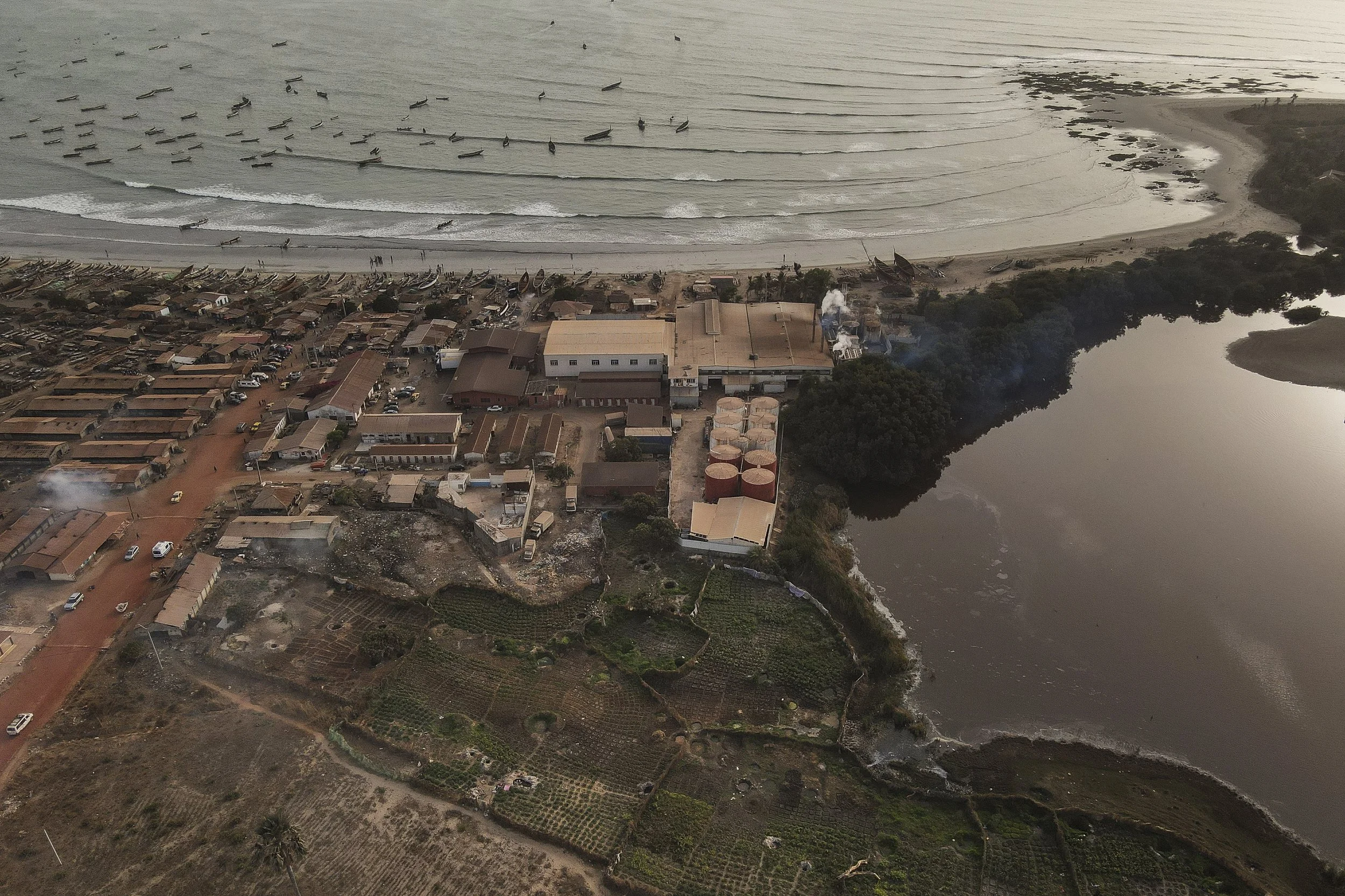 Vista aérea de un pueblo costero con casas, caminos de tierra, un río y una playa con olas, donde se observan barcos en el mar y algunas áreas de cultivo en tierra.