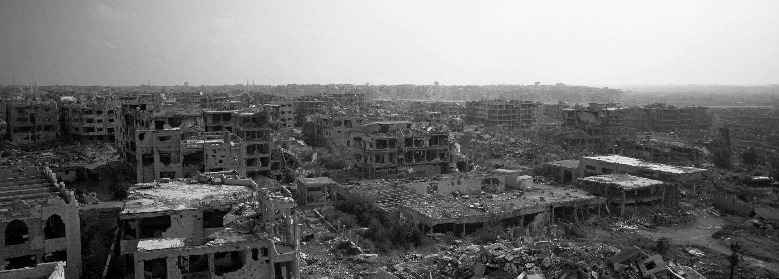 Paisaje urbano de edificios dañados y destruídos en un entorno de guerra o desastre natural, en escala amplia y en tonos grises.