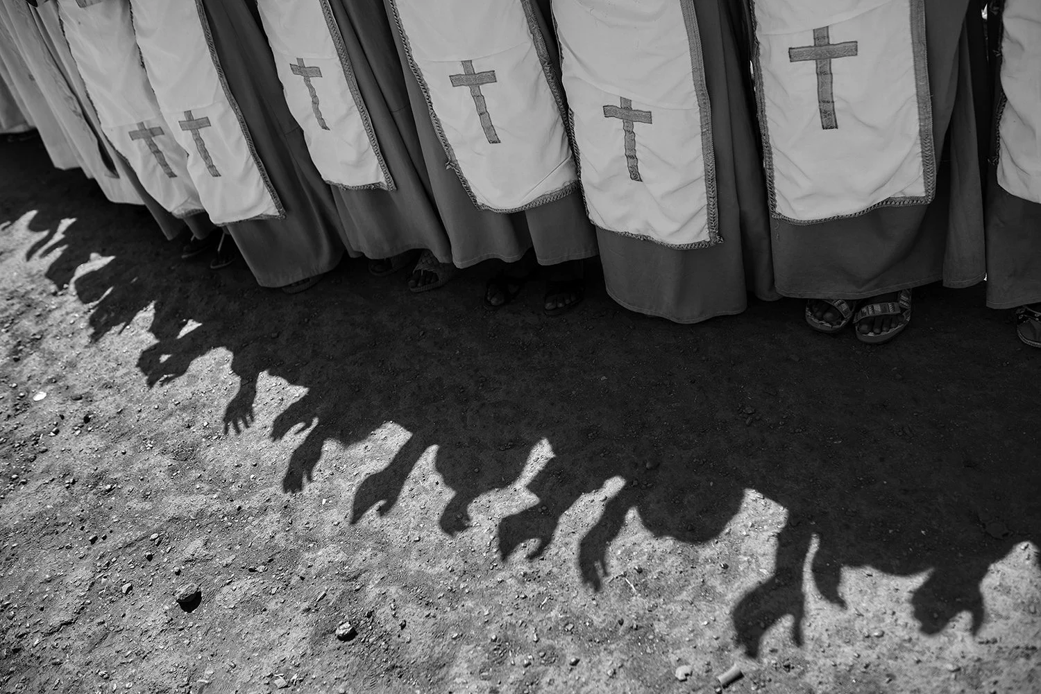 Personas vestidas con túnicas negras y capas blancas con cruces, mirando hacia abajo, con sombras en el suelo que parecen manos de animales o aliens.