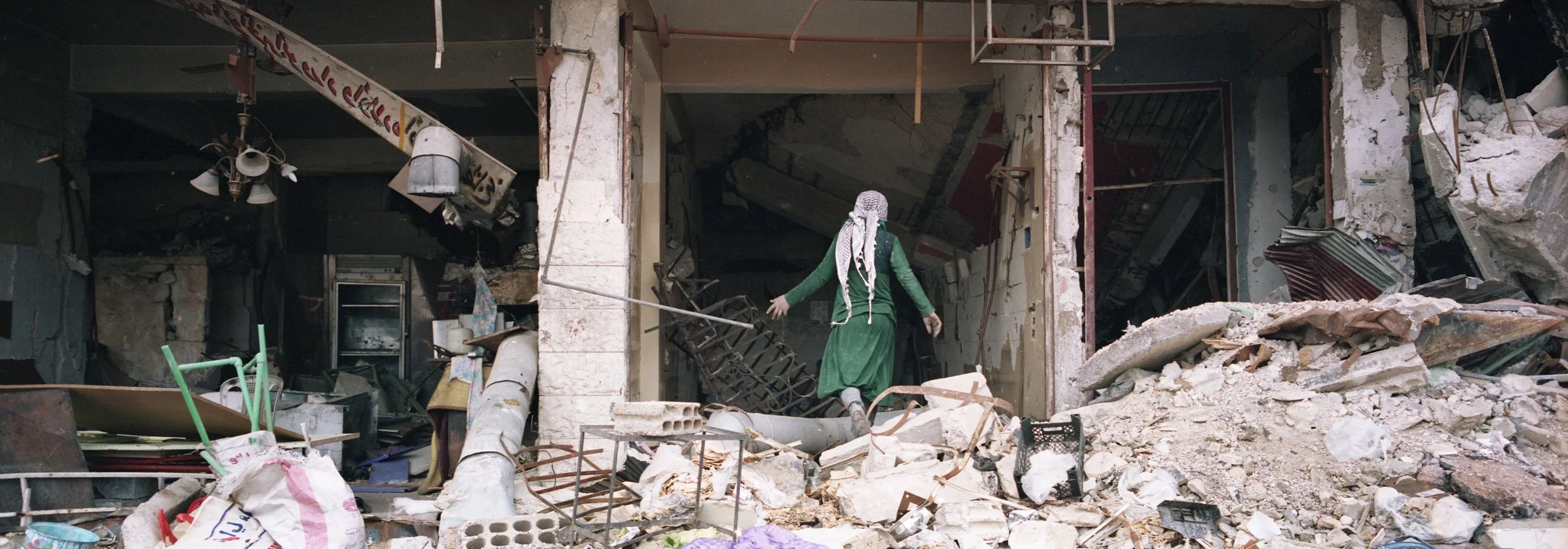 Persona con ropa verde y pañuelo en la cabeza caminando entre escombros tras un desastre
