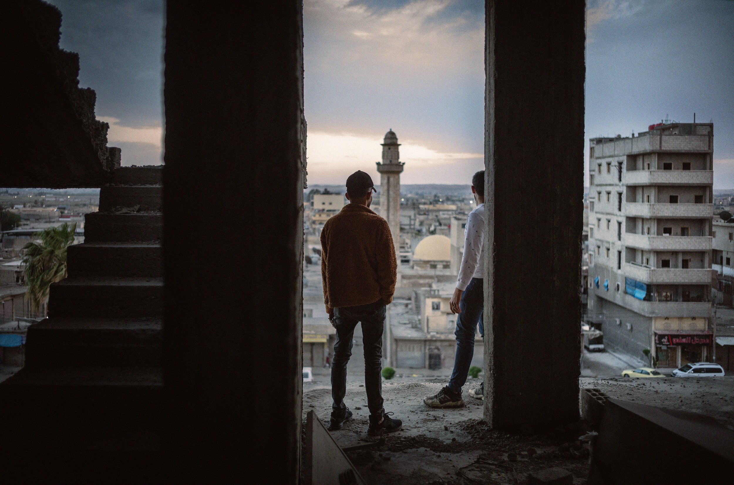 Dos personas de espaldas conversando en una construcción en el ático, con vista a una ciudad y un minarete al fondo, durante el atardecer.