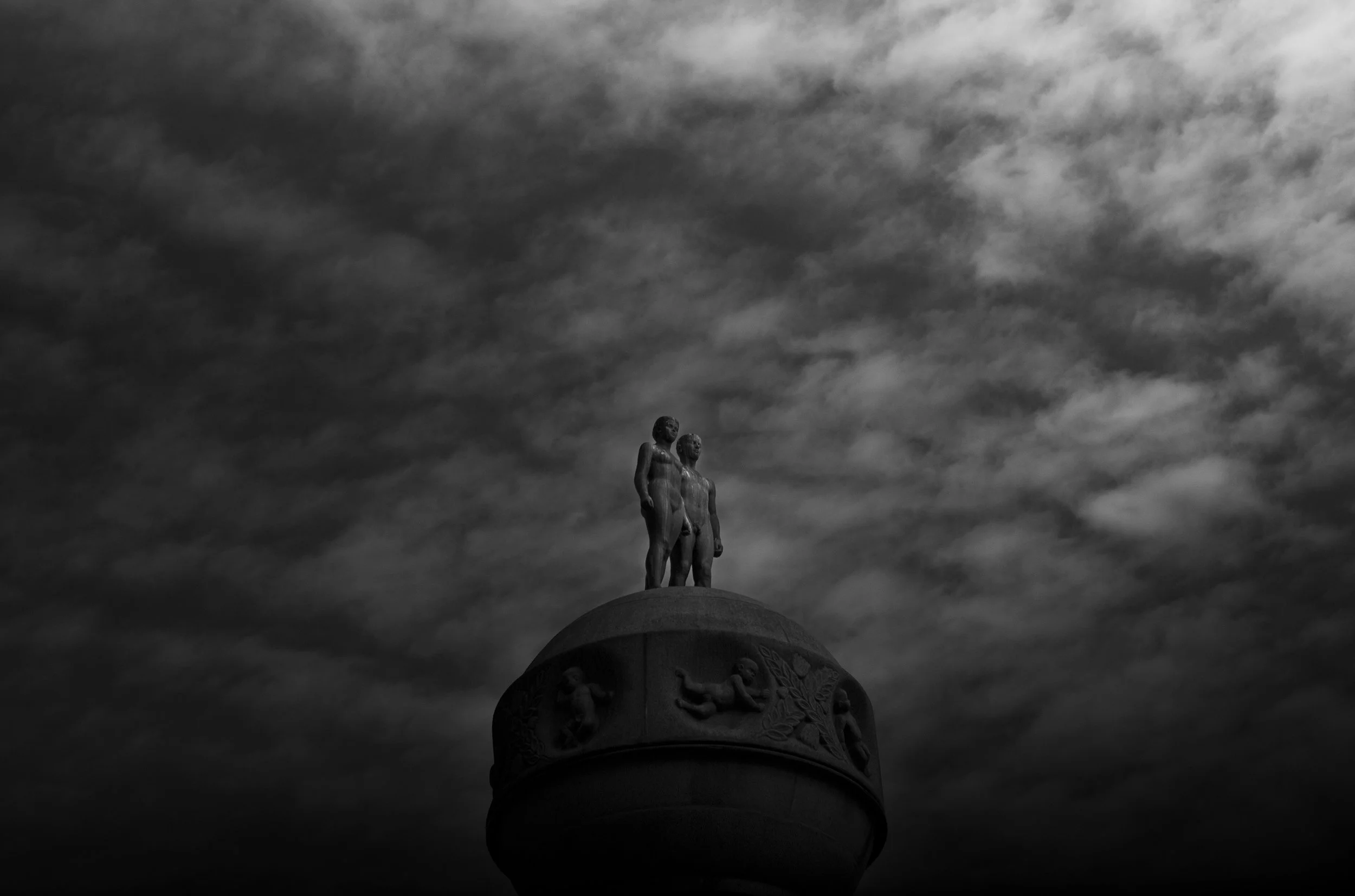 Estatua de dos figuras humanas en la cima de un monumento con relieves, bajo un cielo nublado en tonos oscuros.
