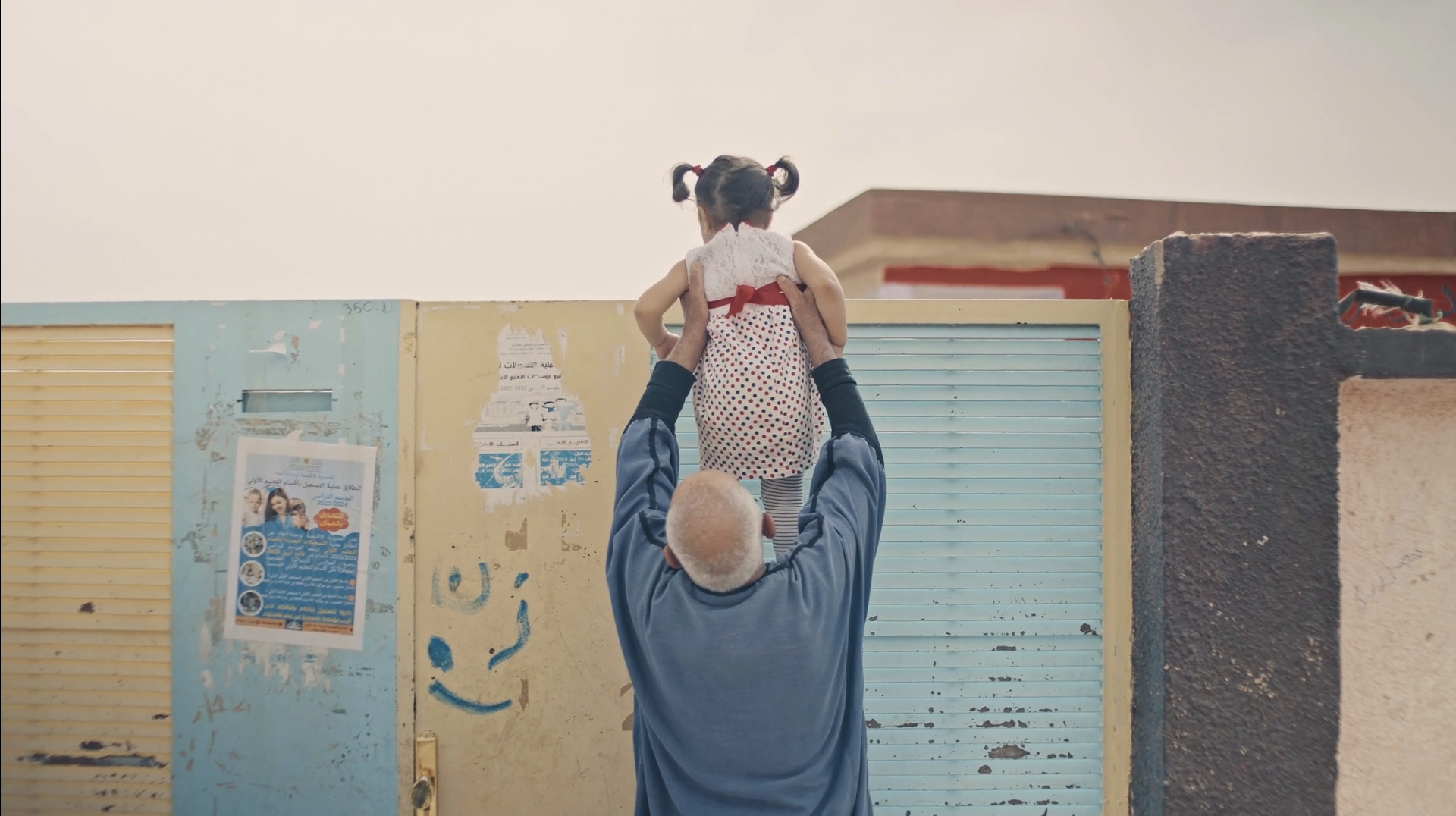 Un anciano con cabello canoso sostiene en alto a una niña con coletas y vestido de puntos blancos con rojo, frente a una pared con posters y graffiti.