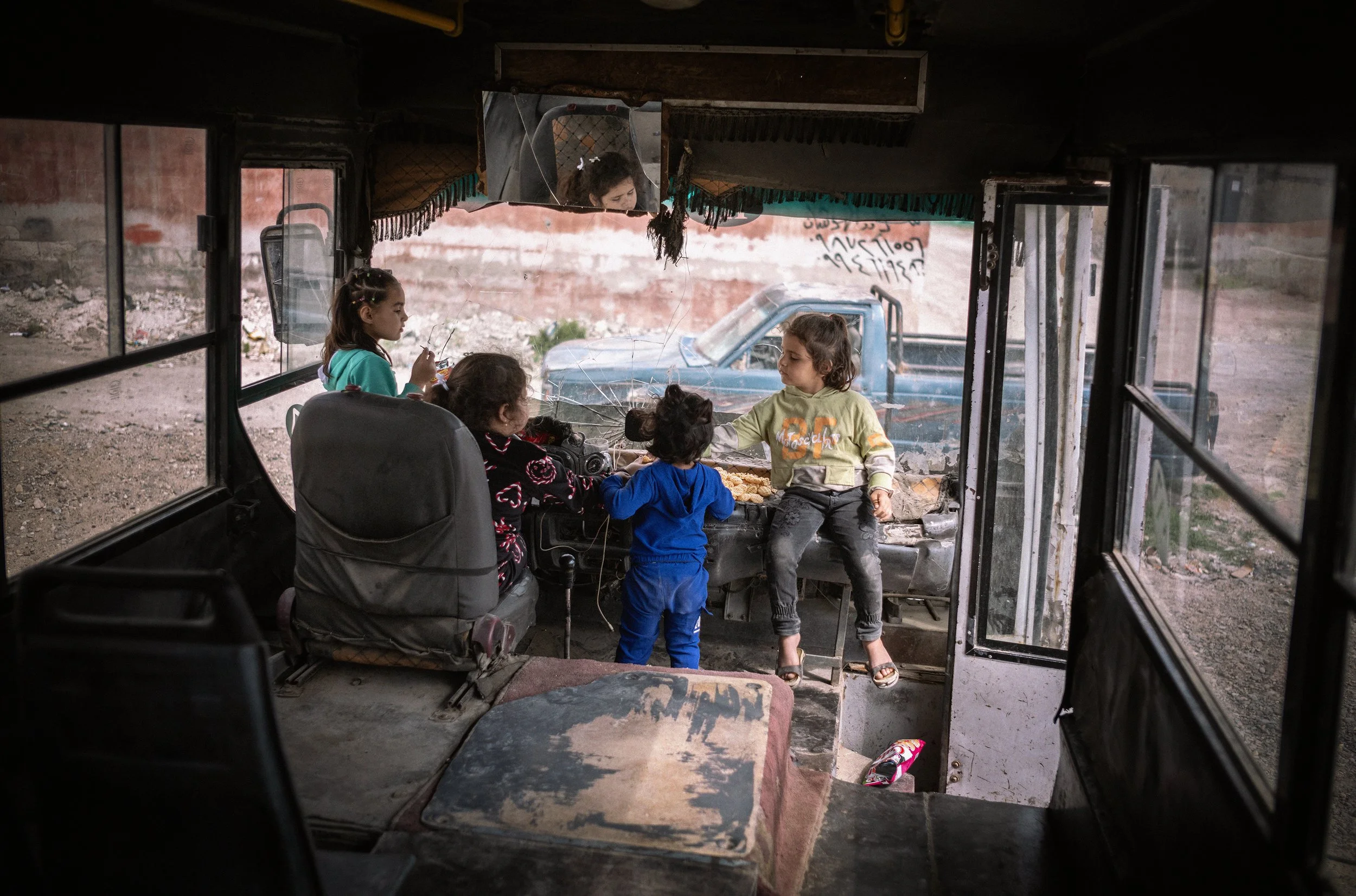 Niños dentro de un autobús en un entorno rural, algunos sentados y otros de pie, con una camioneta visible a través del parabrisas y el vidrio roto del autobús.