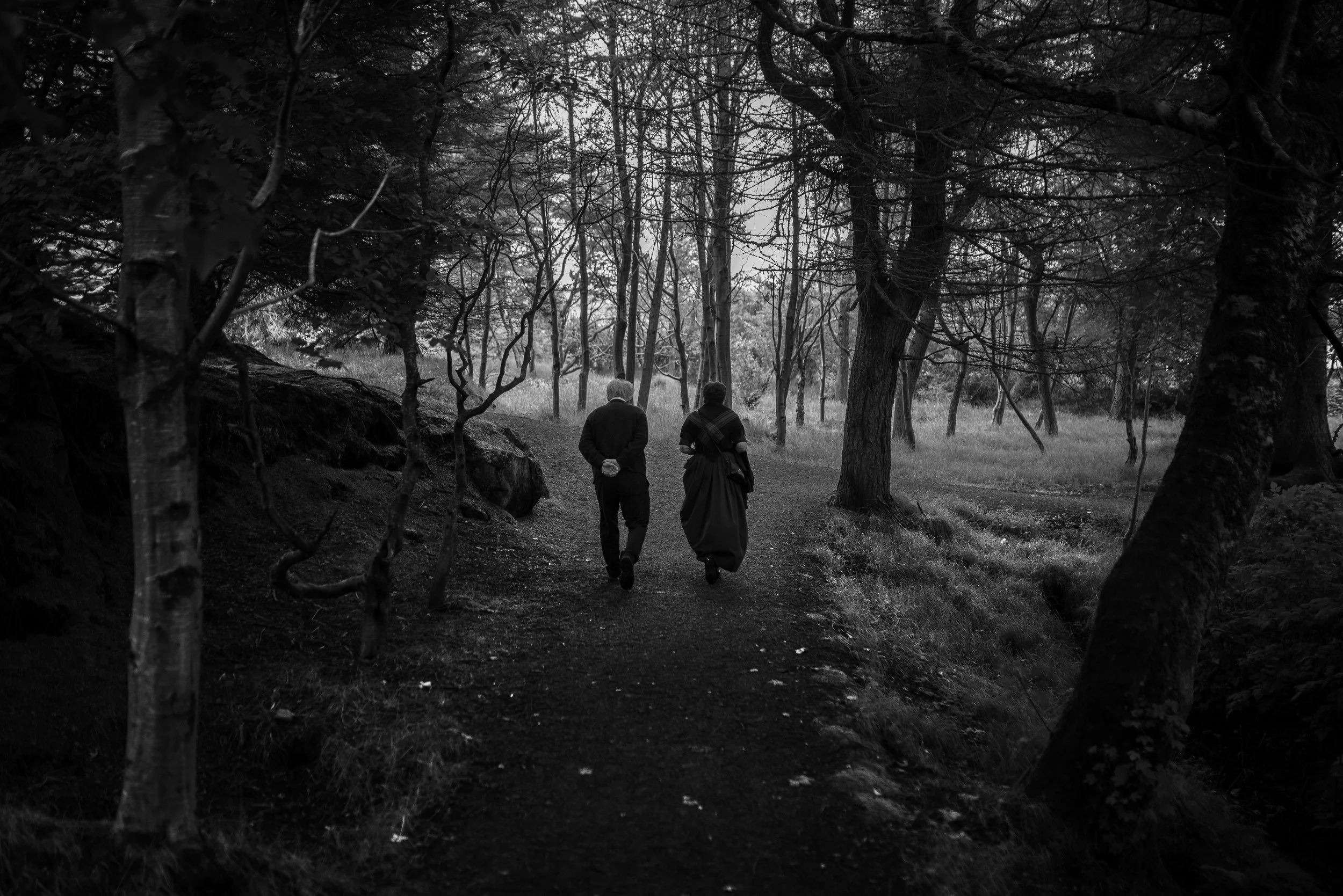 Dos personas caminan por un sendero en un bosque en blanco y negro, rodeadas de árboles y vegetación.