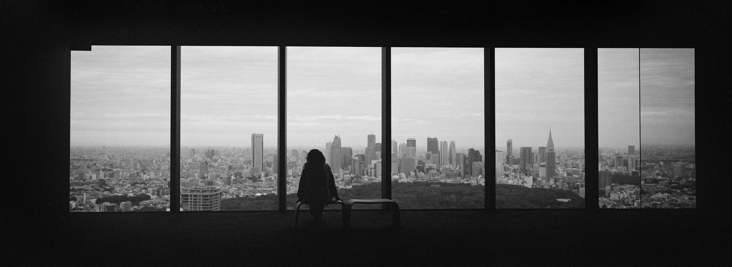 Persona en silueta mirando un horizonte de ciudad desde una ventana grande, en un ambiente oscuro.