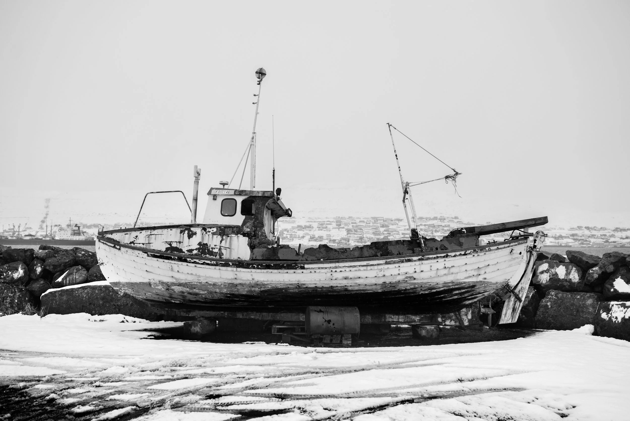 Barco viejo en la orilla, rodeado de piedras y nieve, en un fondo de ciudad y barcos en el mar.