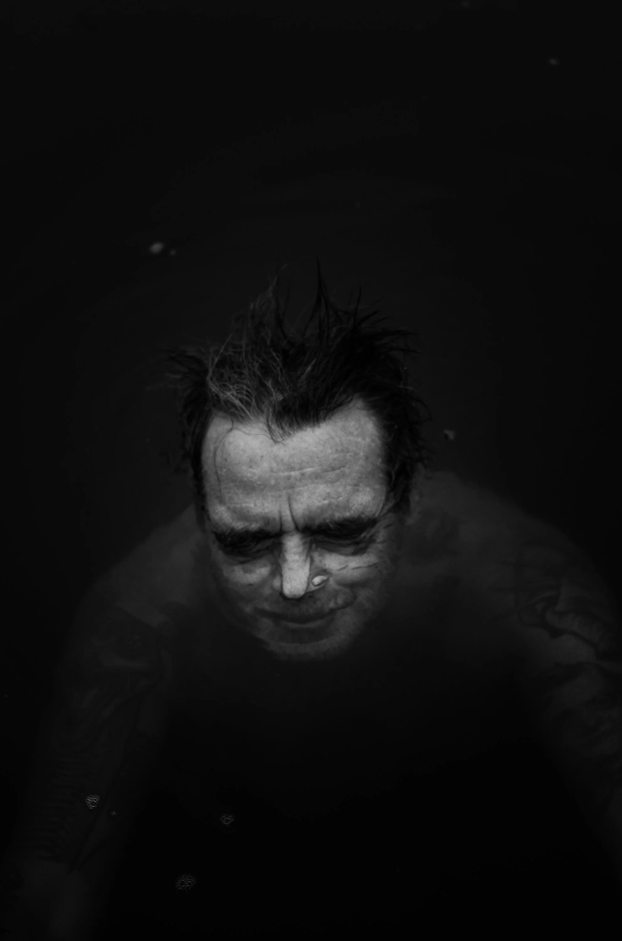 Hombre con cabello mojado o arreglado de manera desordenada, sumergido en agua oscura, vista desde arriba.