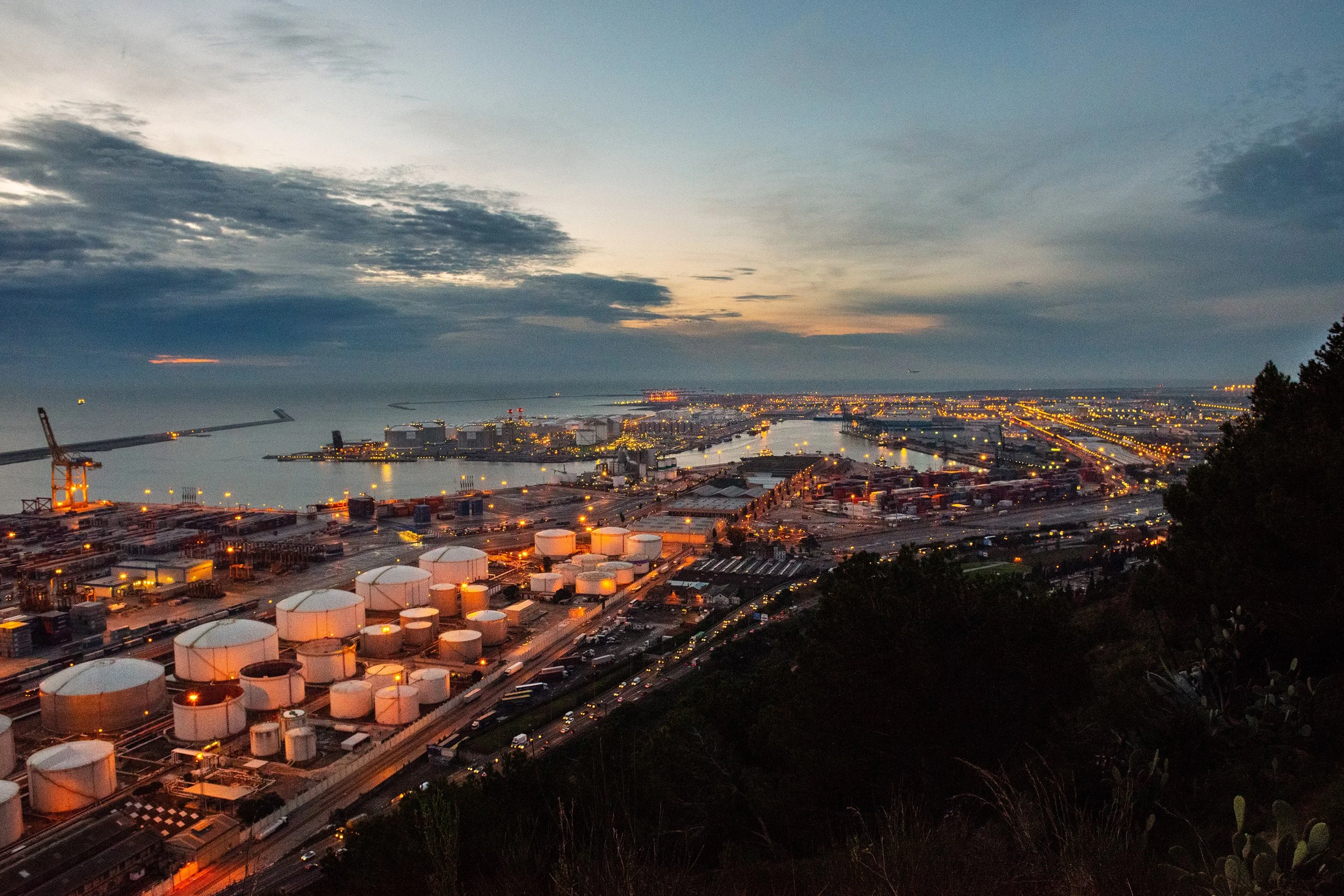 Vista panorámica de un puerto industrial al atardecer con tanques de almacenamiento, grúas y vías de tráfico.
