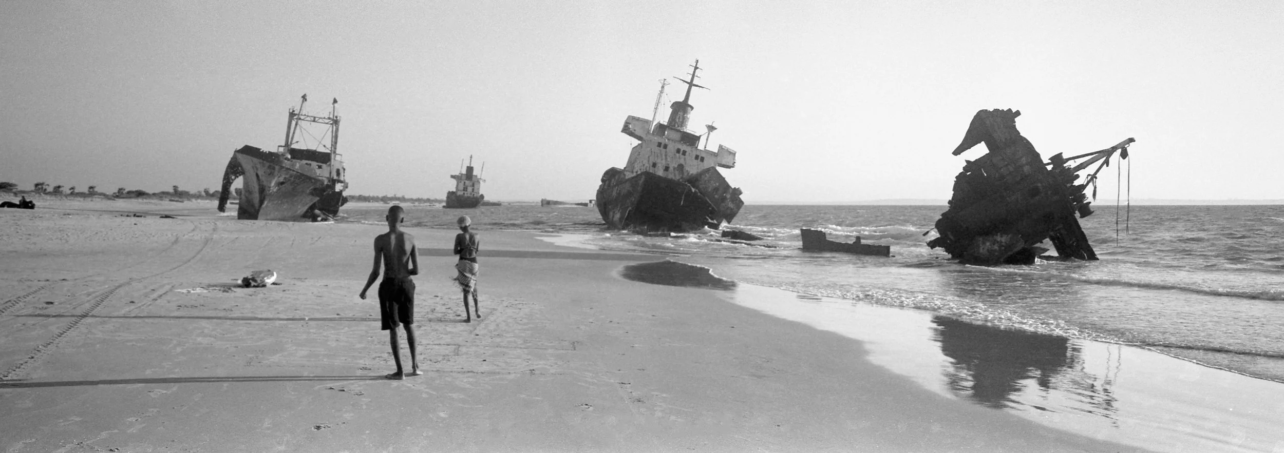 Navíos varados en la orilla de una playa de arena, con dos personas caminando en primer plano en un día despejado.