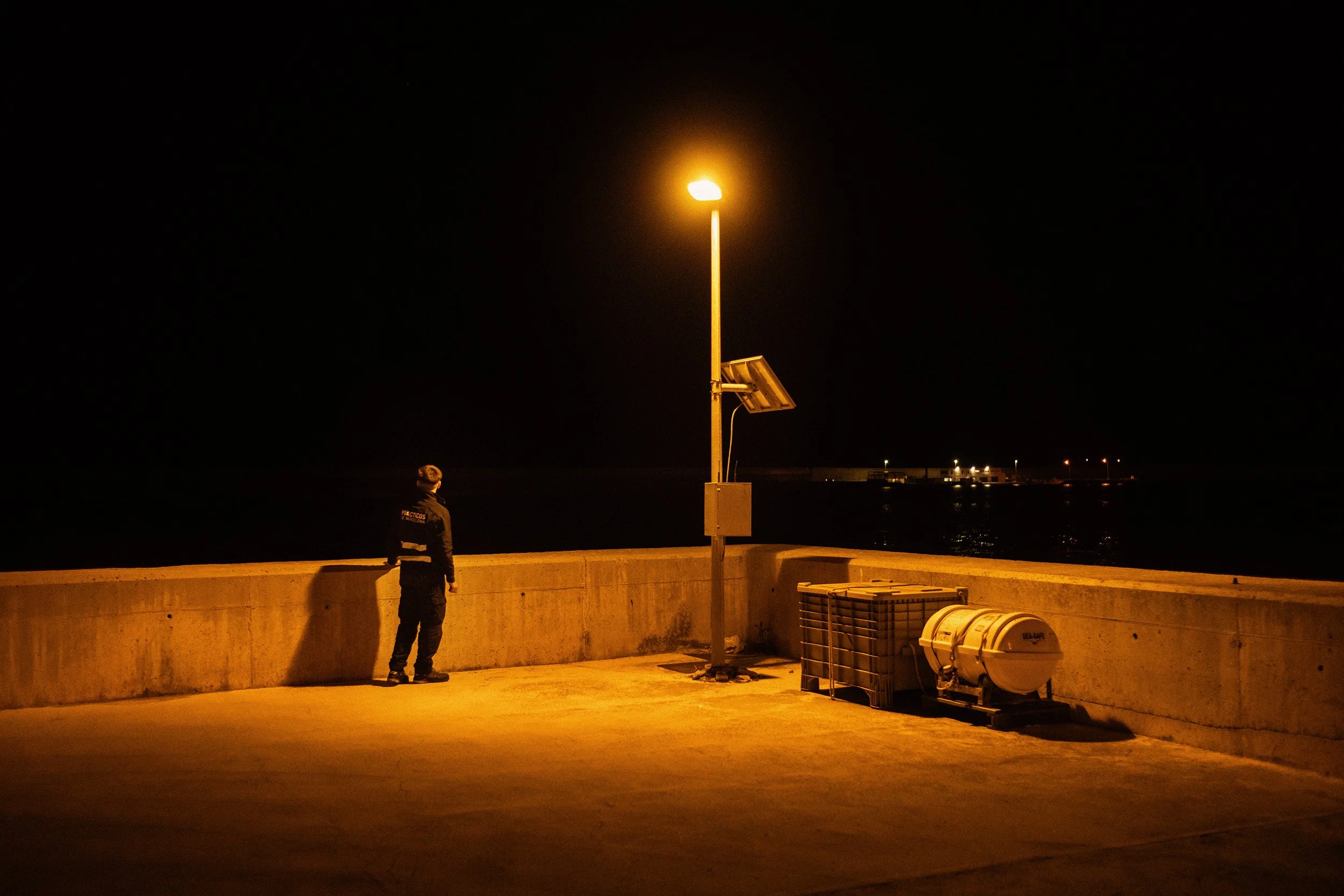 Persona de espaldas en un muelle en la noche, iluminado por una farola, con objetos como barriles y una caja en el suelo, y un mar en el fondo.