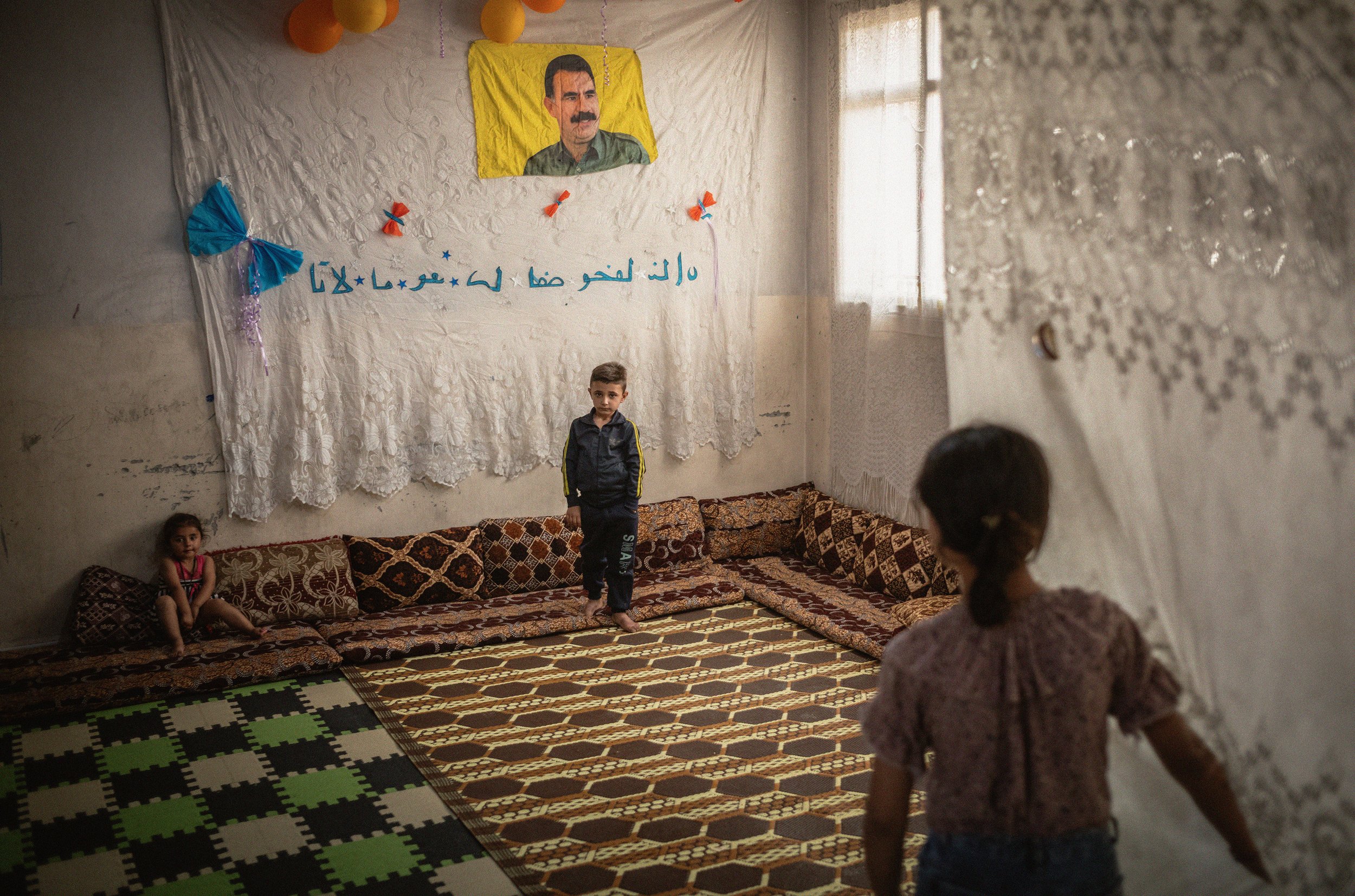 Una habitación decorada con un retrato grande y una bandera, con niños y una mujer en el interior. El fondo tiene cortinas blancas y un tapiz en la pared con escritura en árabe.