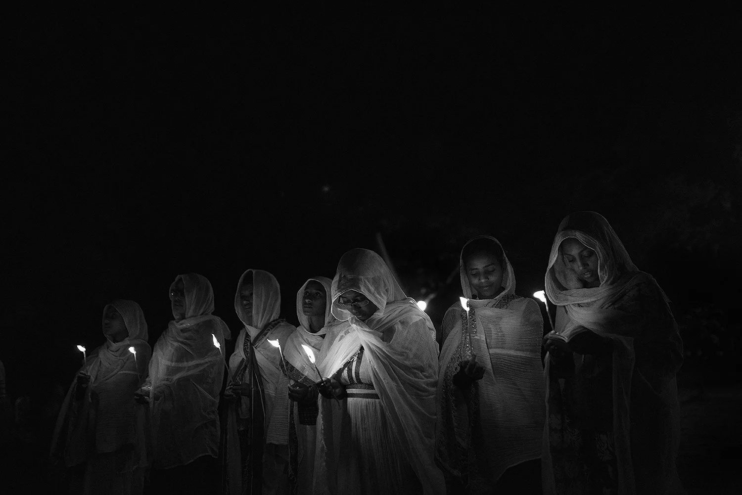 Grupo de mujeres vestidas con ropa tradicional, sosteniendo velas encendidas en la noche, con fondo oscuro