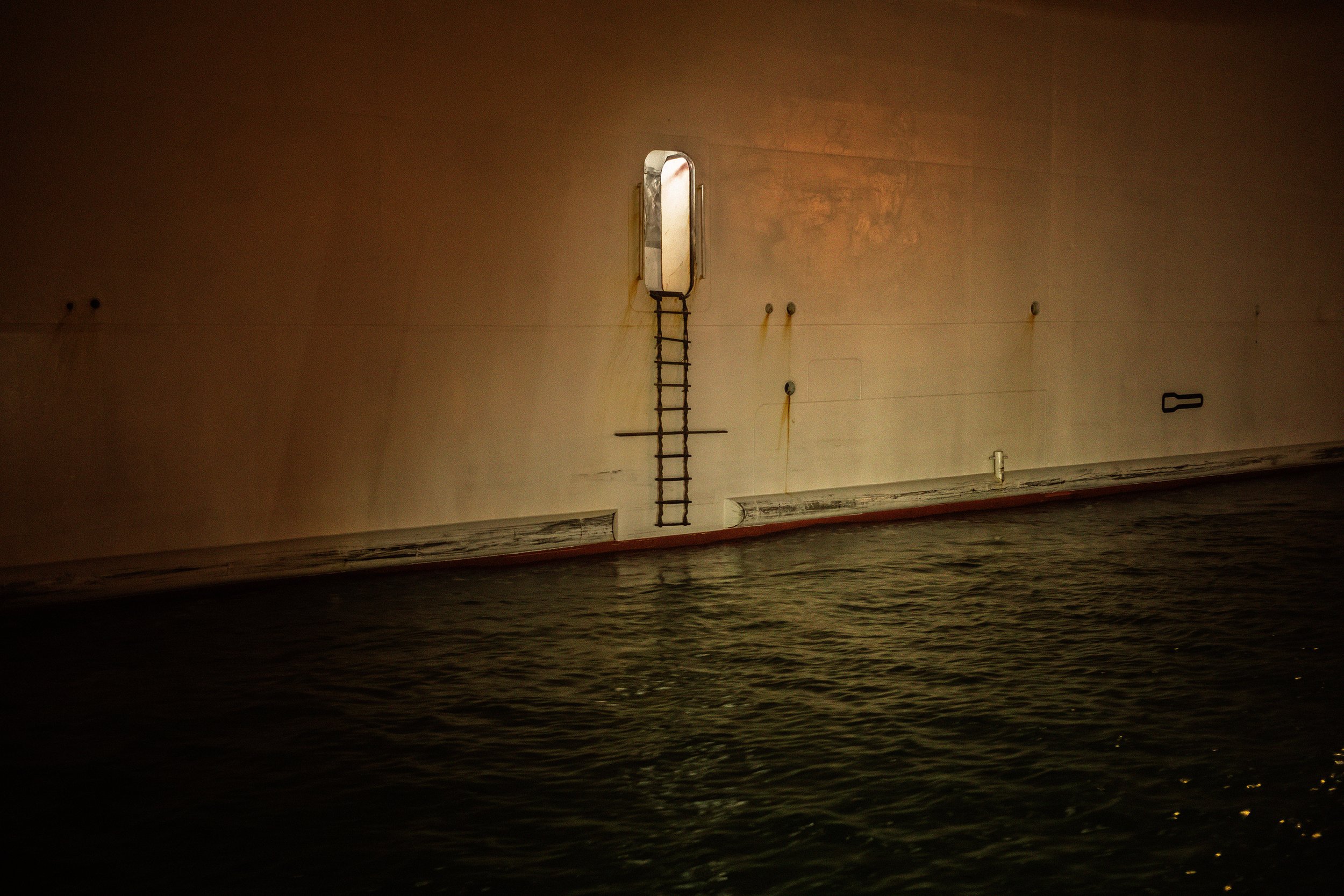 Vista de un barco desde el agua en la noche, mostrando la plancha lateral del navío con una escalera y una ventana en la parte superior