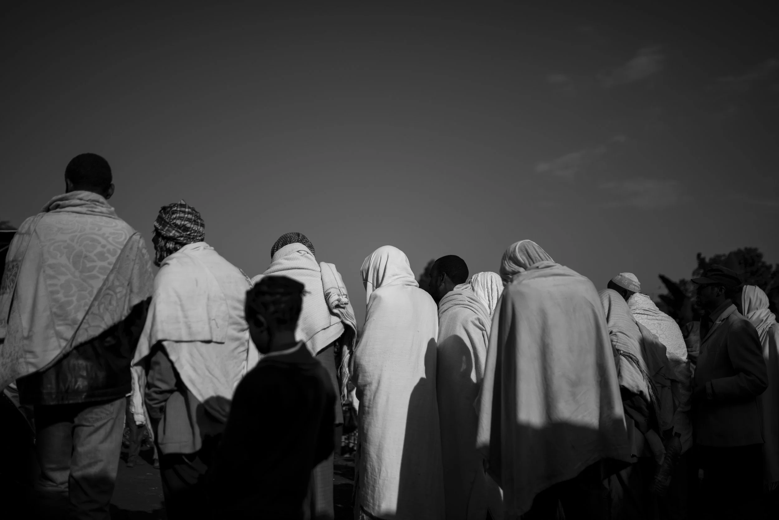 Grupo de personas con ropas tradicionales, en línea, en un espacio abierto bajo cielo despejado, en negro y blanco.