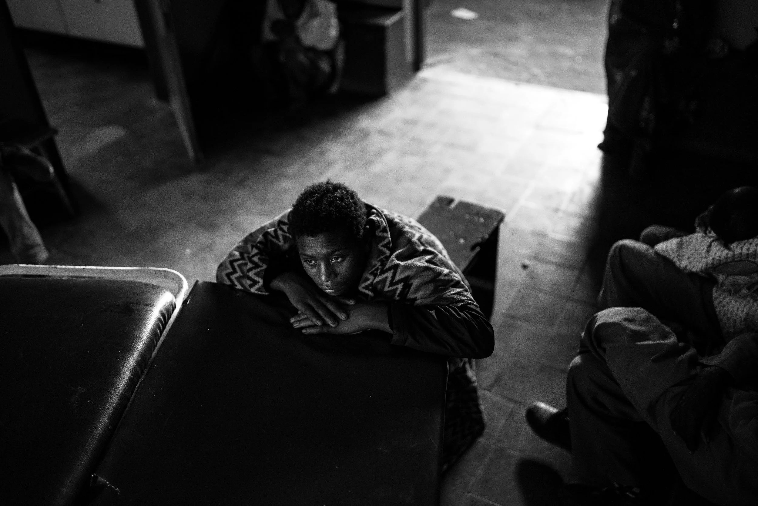 Joven sentado en un establecimiento, apoya la cabeza en sus brazos sobre una mesa, en un ambiente oscuro y con luz que entra por la puerta abierta.