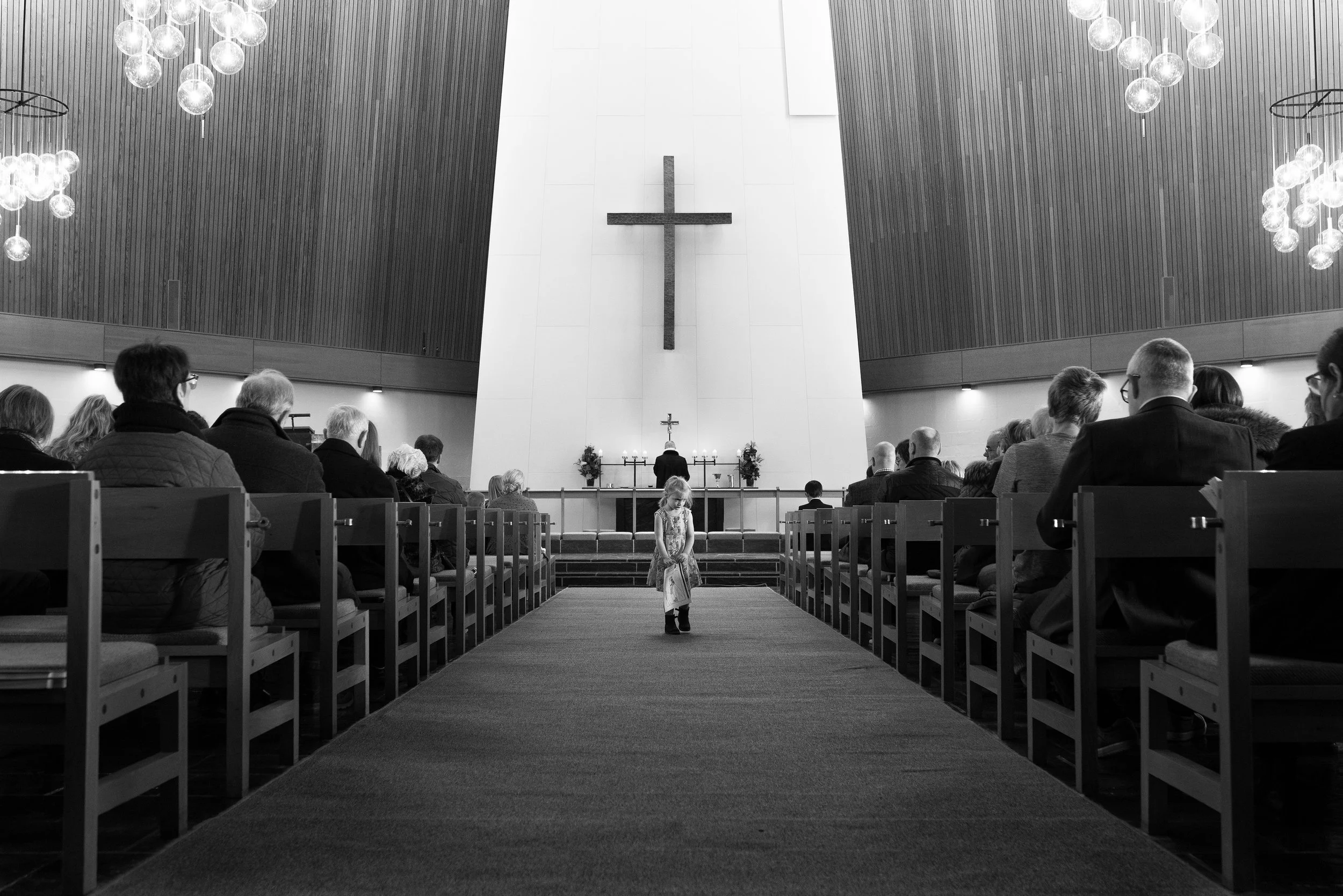 Interior de una iglesia con feligreses sentados en bancas, una niña en el pasillo central, al fondo un altar con una cruz grande en la pared y un sacerdote de espaldas.