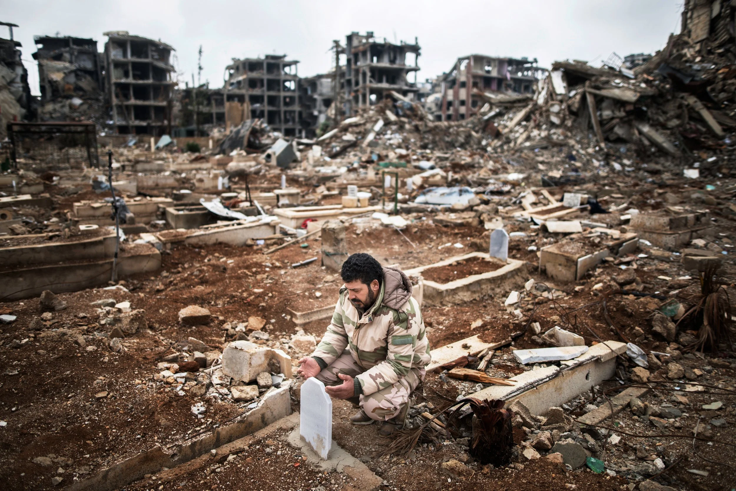 Hombre con uniforme militar en un cementerio destruido, orando o recordando a los fallecidos en un escenario de desastre o guerra.