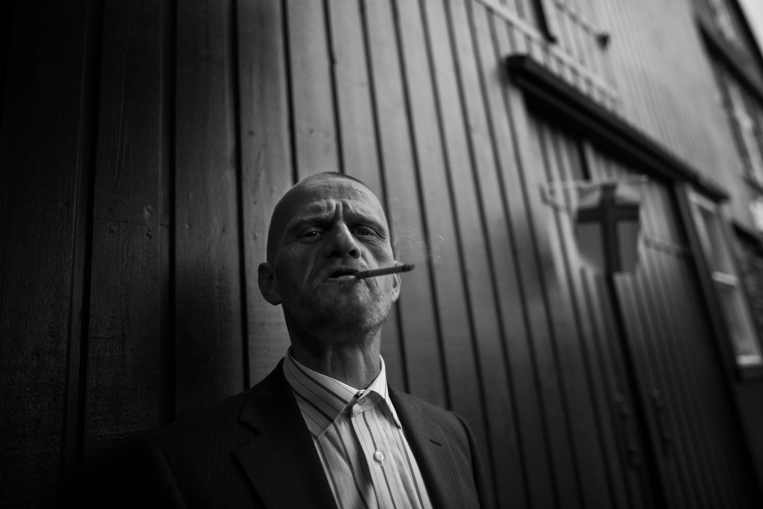 Un hombre con una camisa de rayas, chaqueta y sin cabello, fuma un cigarrillo y mira hacia la cámara, de fondo una pared de madera oscura con ventanas