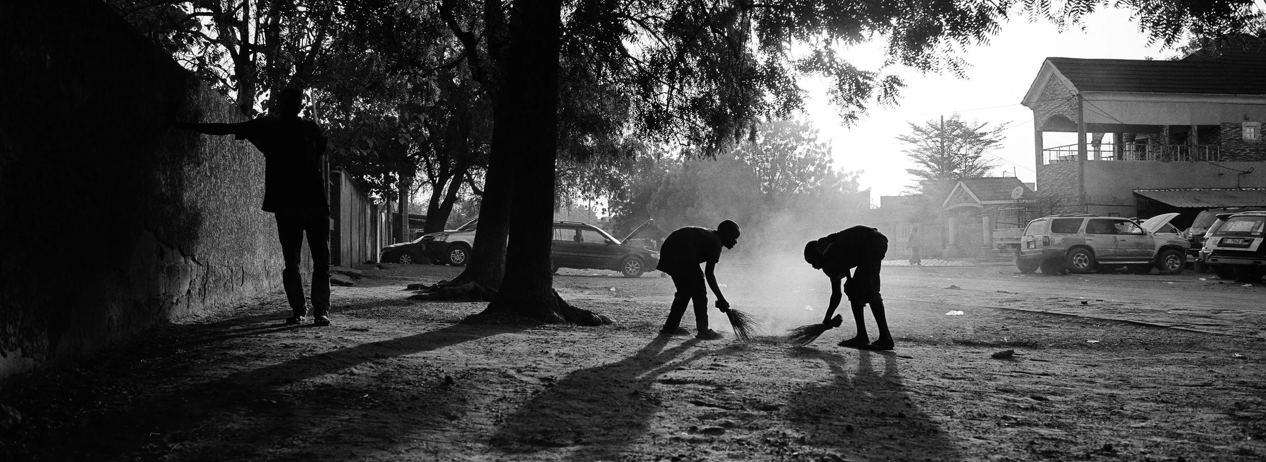 Tres personas barriendo en una calle polvorienta al atardecer, con varios autos estacionados y árboles en el fondo en un entorno urbano.