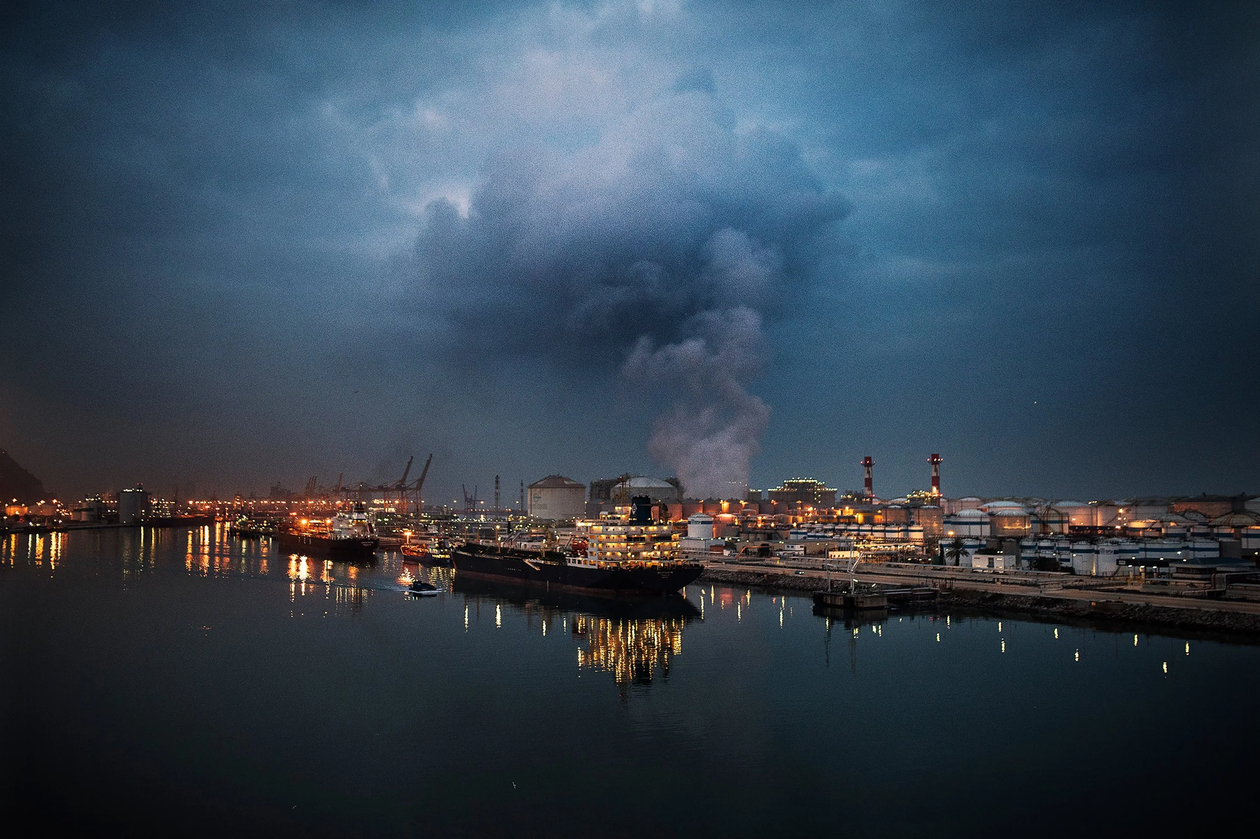 Vista nocturna de un puerto industrial con barcos y naves, columnas de humo y una gran nube en el cielo