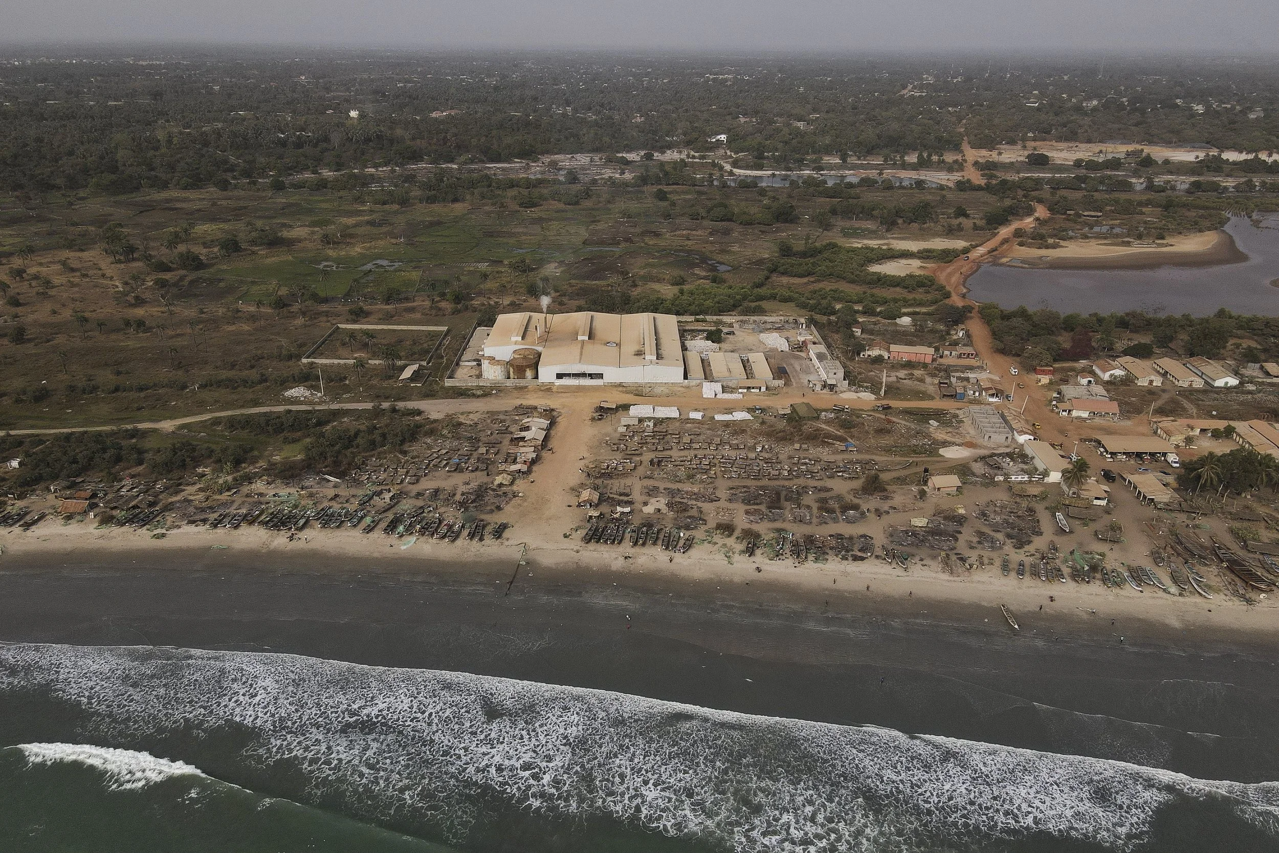 Vista aérea de una playa con muchas motos y carros en la arena, un área industrial y caminos rurales rodeados de vegetación y cuerpos de agua en el fondo.