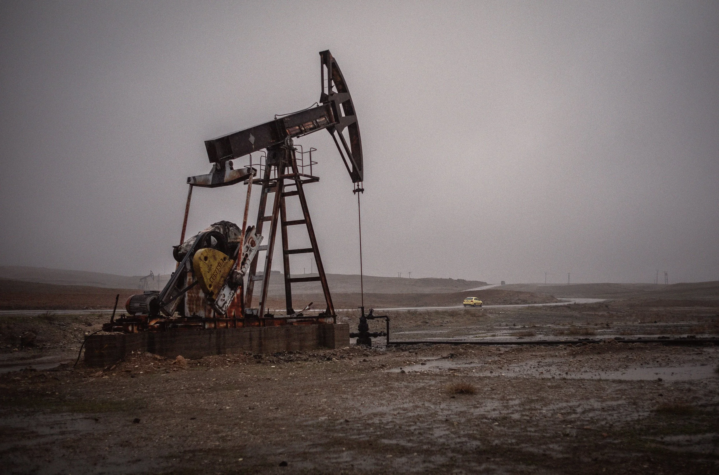 Poza de un pozo petrolero en un paisaje árido con cielo nublado y una camioneta amarilla en la distancia.