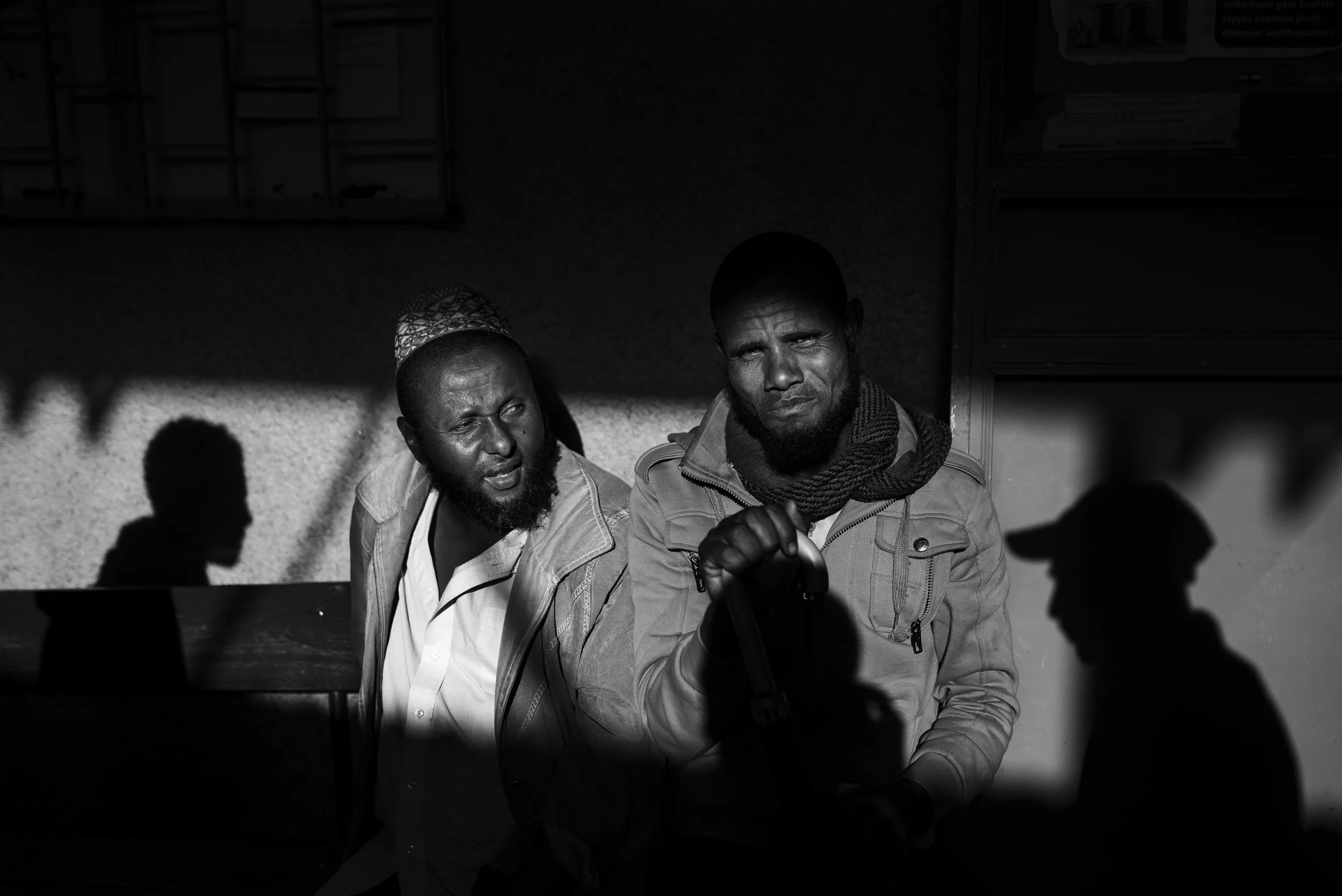 Dos hombres con barba y sin barba en una sombra, uno mirando a la cámara y otro conversando, en un entorno exterior en blanco y negro.