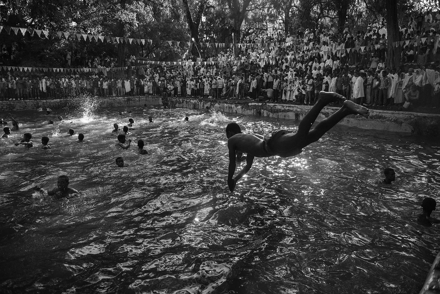 Muchos niños jugando y nadando en una piscina llena de agua, con una multitud de personas observando desde la orilla y árboles en el fondo, en un evento al aire libre.
