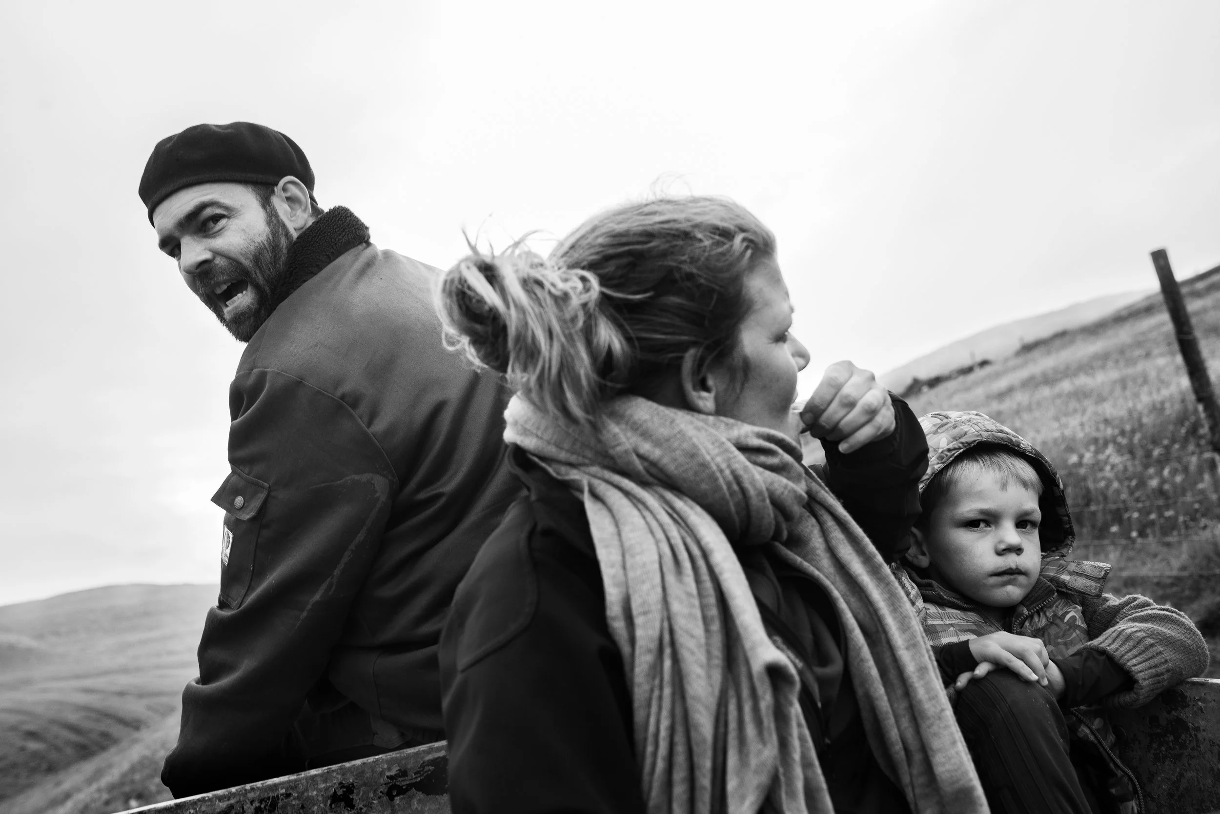 Una familia viajando en una carroza en un campo con cielo nublado, incluyendo un hombre con barba y gorra, una mujer con bufanda y un niño con abrigo, en una vista en blanco y negro.