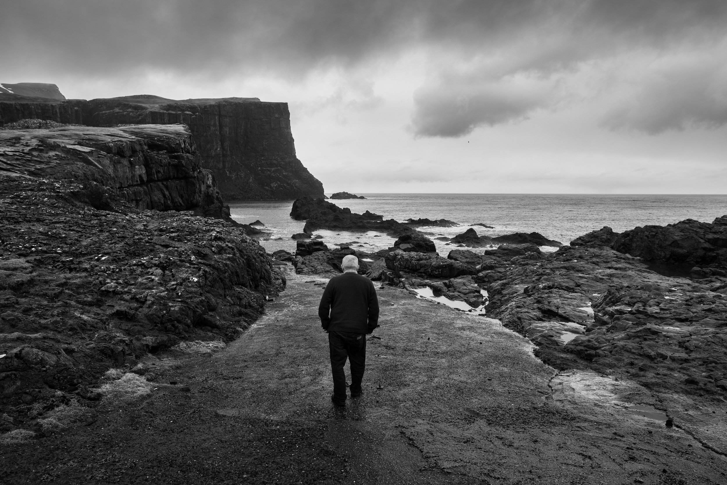 Un hombre caminando en un sendero junto al mar, con acantilados en el fondo, en un día nublado en un paisaje costero en tonos de gris.