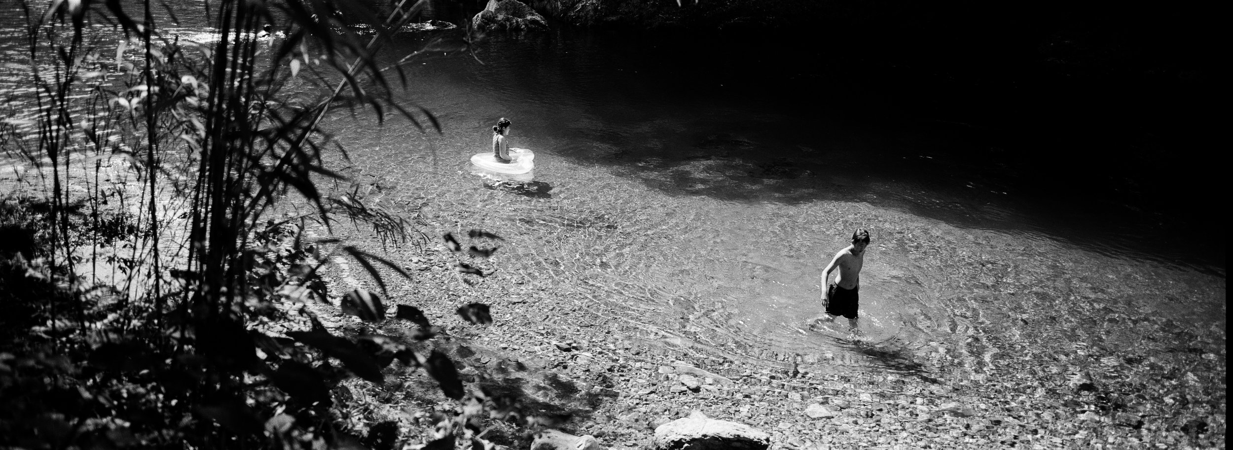 Dos niños en un río o lago con agua clara, uno de ellos en el agua y el otro en la orilla, rodeados de vegetación en un entorno natural, en blanco y negro.