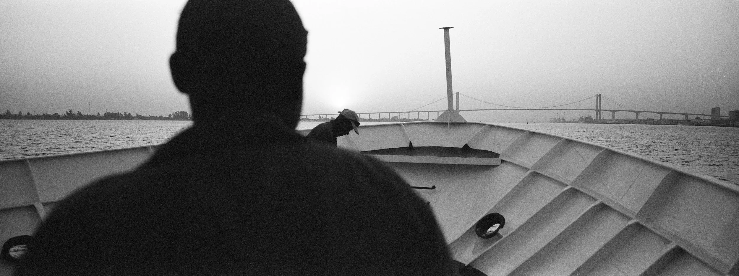 Fotografía en blanco y negro de dos personas en un barco en un cuerpo de agua, con un puente visible en la distancia.