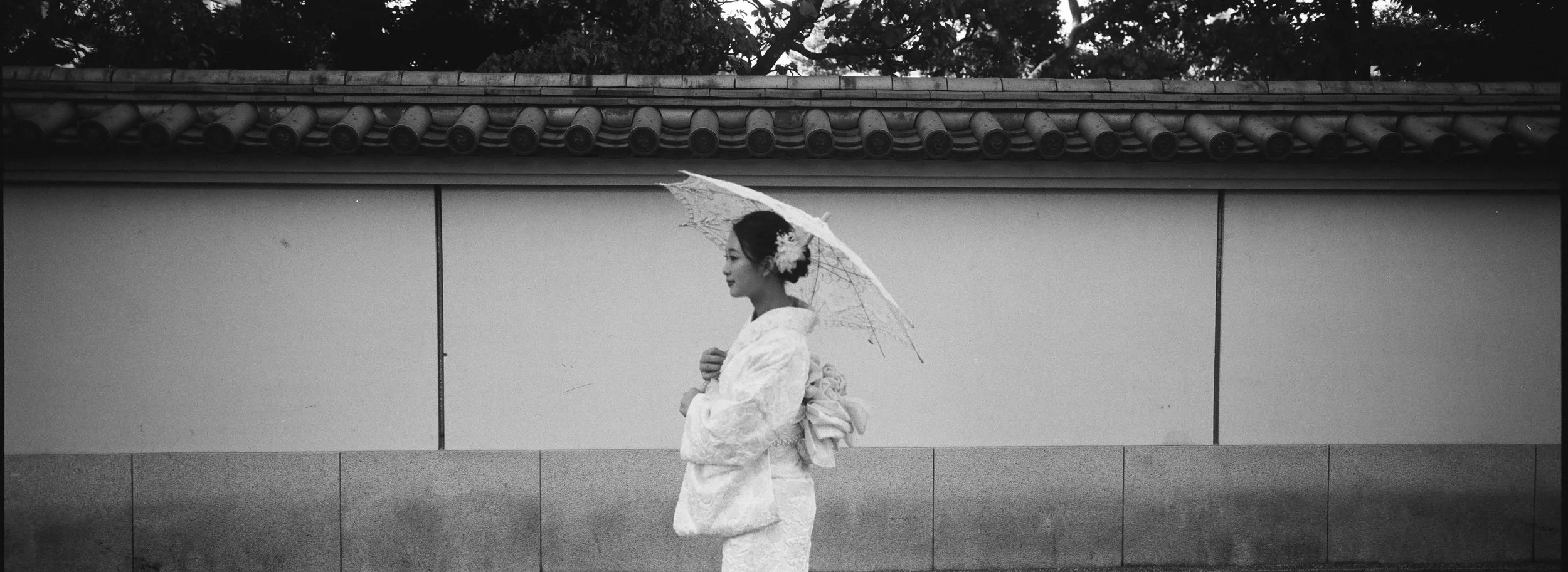 Mujer vestida con kimono tradicional japonés, sosteniendo un paraguas y adornada con flores en el cabello, en un entorno urbano en blanco y negro.