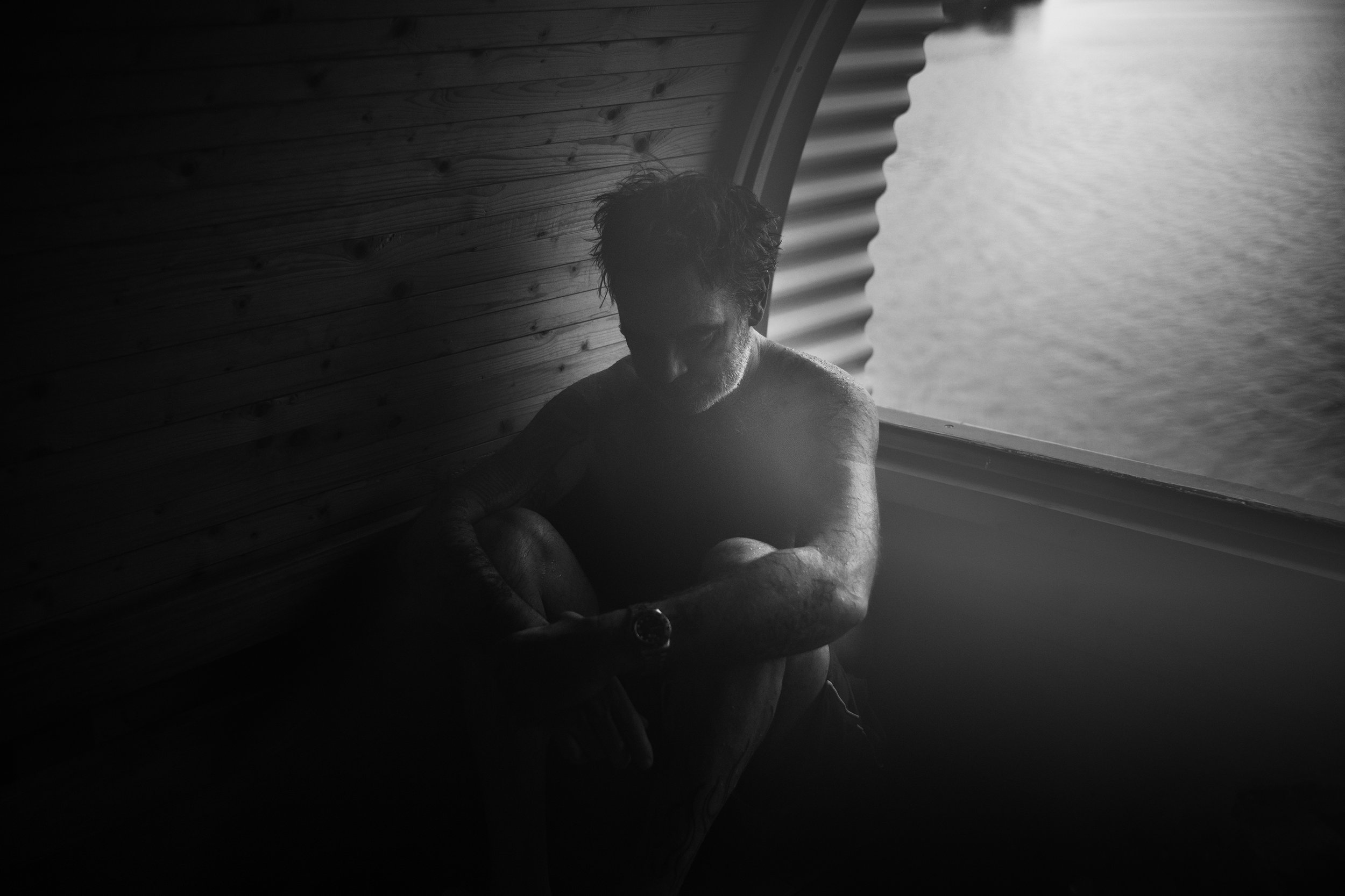 Un hombre con barba y pelo rizado sentado junto a una ventana, en una habitación con paredes de madera, mirando hacia abajo, en un ambiente en tonos oscuros y en blanco y negro.