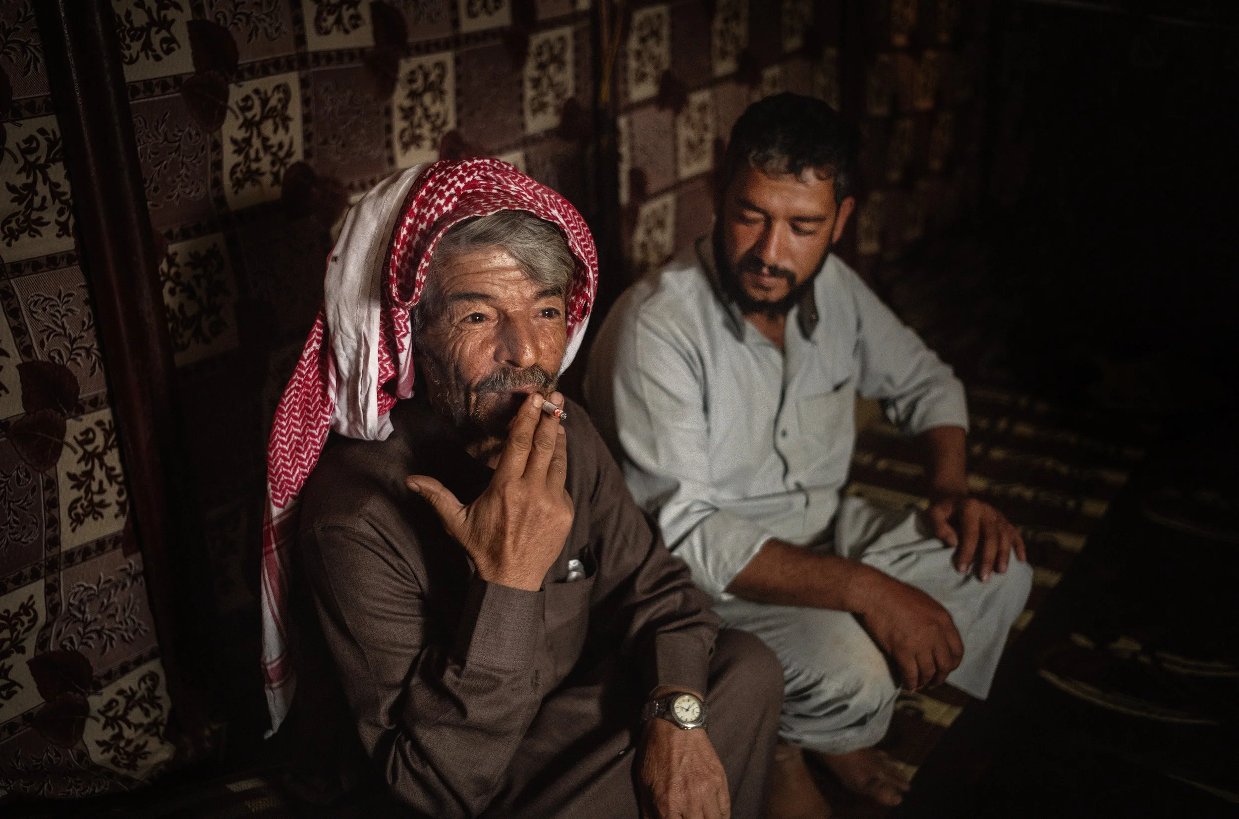 Dos hombres sentados en un ambiente oscuro, uno de ellos con un tocado rojo y blanco, fuma usando un cigarrillo, mientras el otro observa.