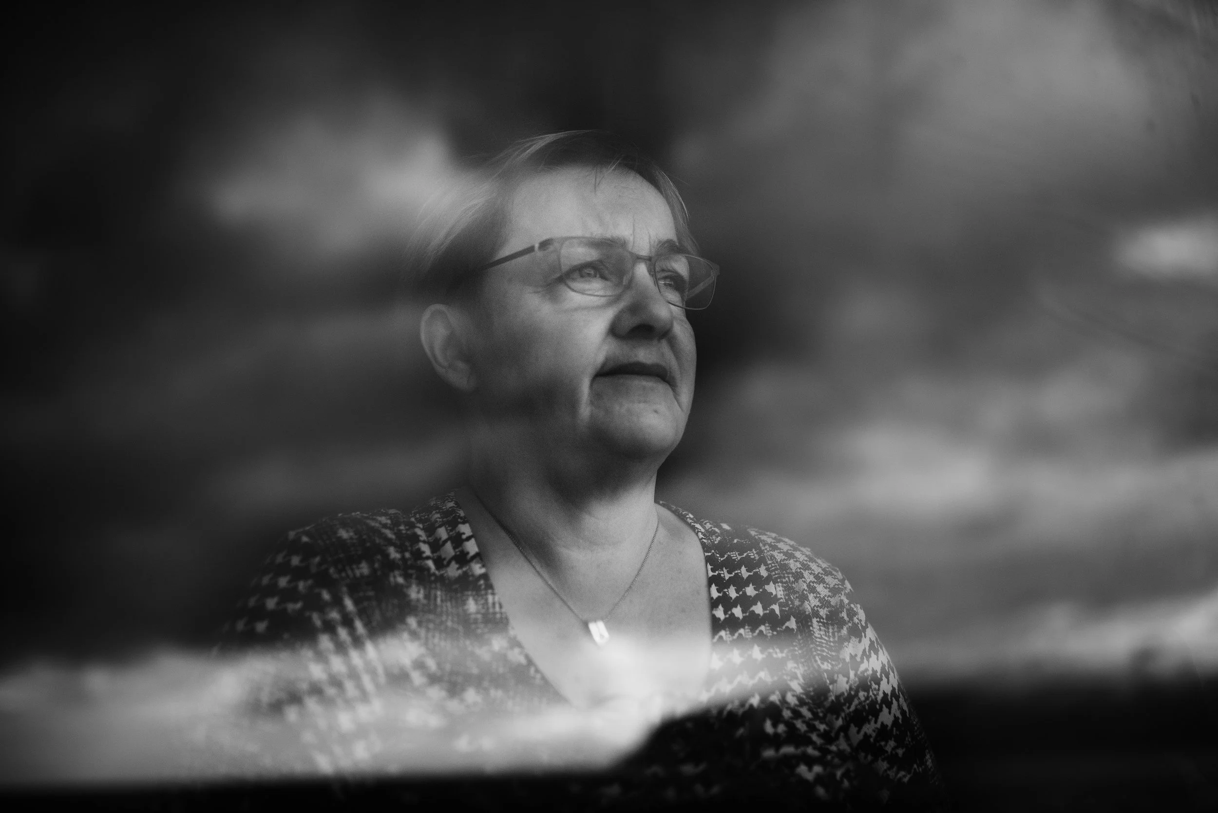 Retrato en blanco y negro de una mujer mayor con gafas, mirando hacia arriba, con un cielo nublado al fondo.