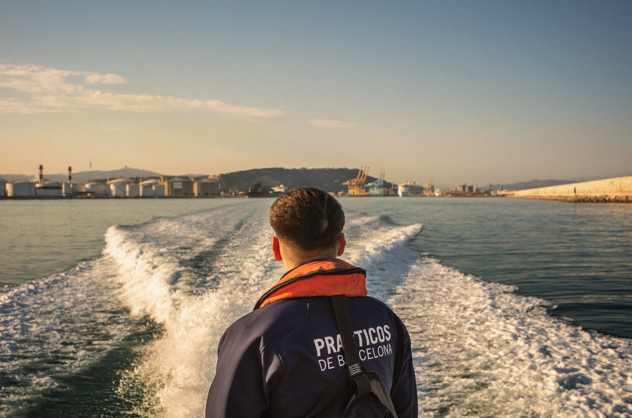 Persona en una lancha en el mar en Barcelona, con vista a puertos y montañas al fondo, durante el día.