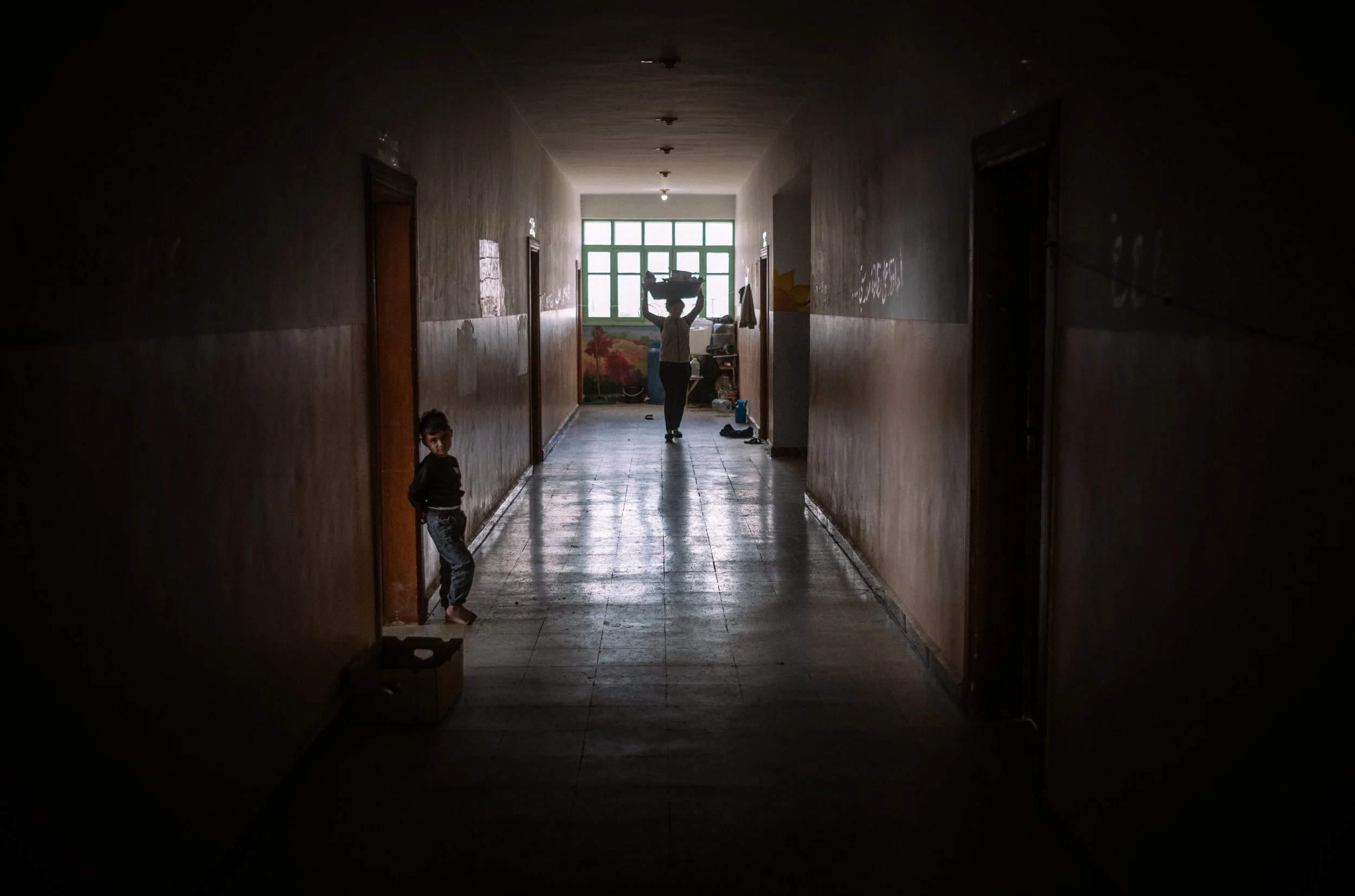 Pasillo largo con paredes de color oscuro, luz natural desde una ventana al fondo, una persona lleva una caja en la cabeza y un niño se asoma desde una puerta a la izquierda.
