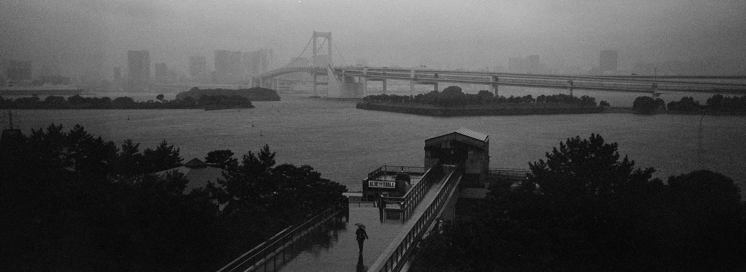 Vista en blanco y negro de un puente y un lago con islas, en un día lluvioso, con una persona con paraguas caminando por un pasillo cubierto a la orilla del lago.