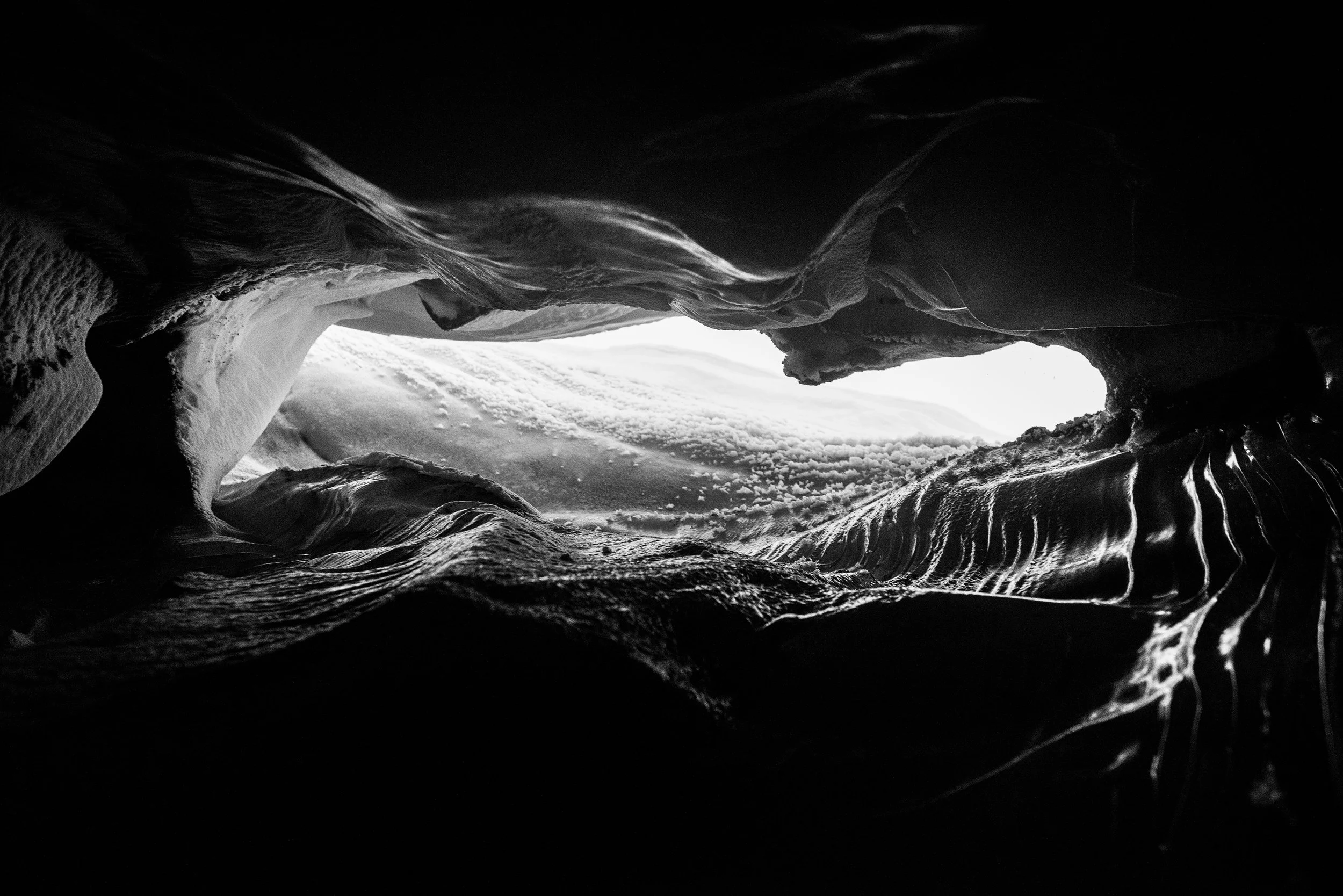 Vista desde adentro de una cueva con la entrada iluminada y formaciones de hielo en las paredes, en tonos negros y blancos.