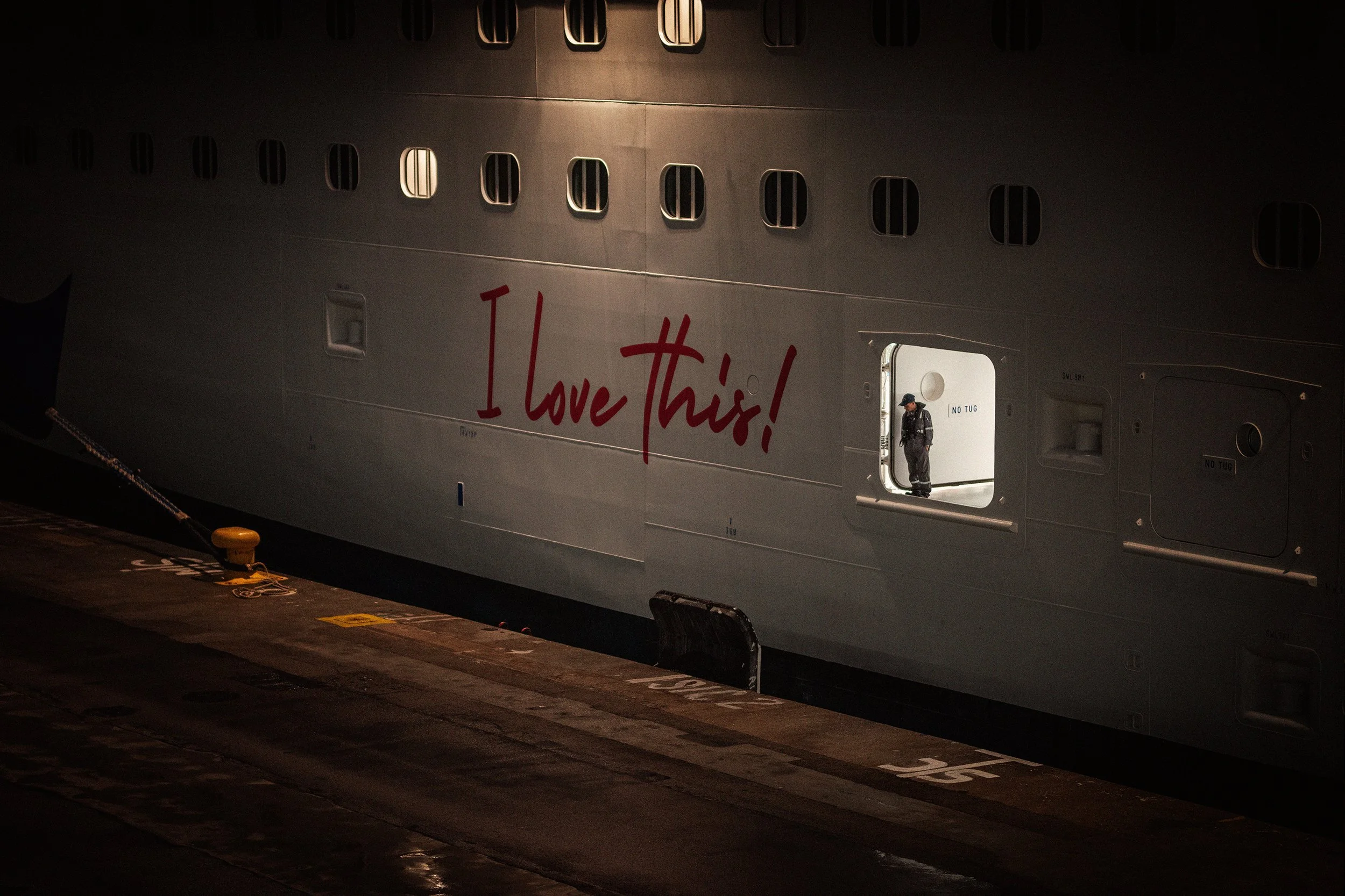 Una nave espacial en un muelle, con la frase 'I love this!' escrita en la parte lateral en letras rojas. Un guardia o trabajador en uniforme está de pie en una escotilla de la nave, visible a través de una abertura. La escena está en la noche o en un