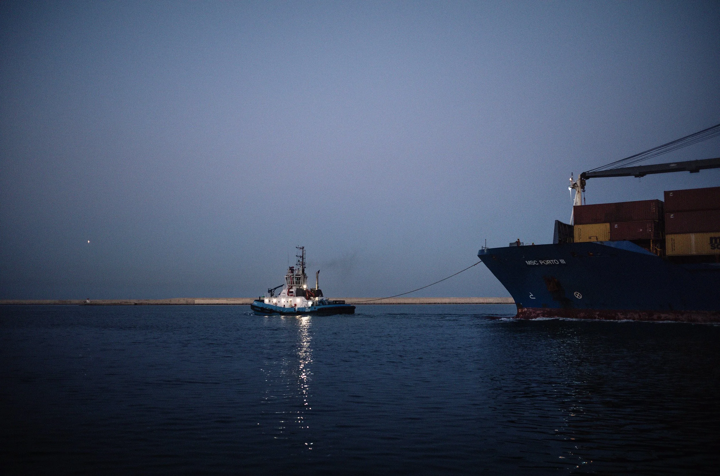 Un barco de carga y una embarcación de remolque en un puerto durante la noche, con el mar y el cielo oscuro de fondo.