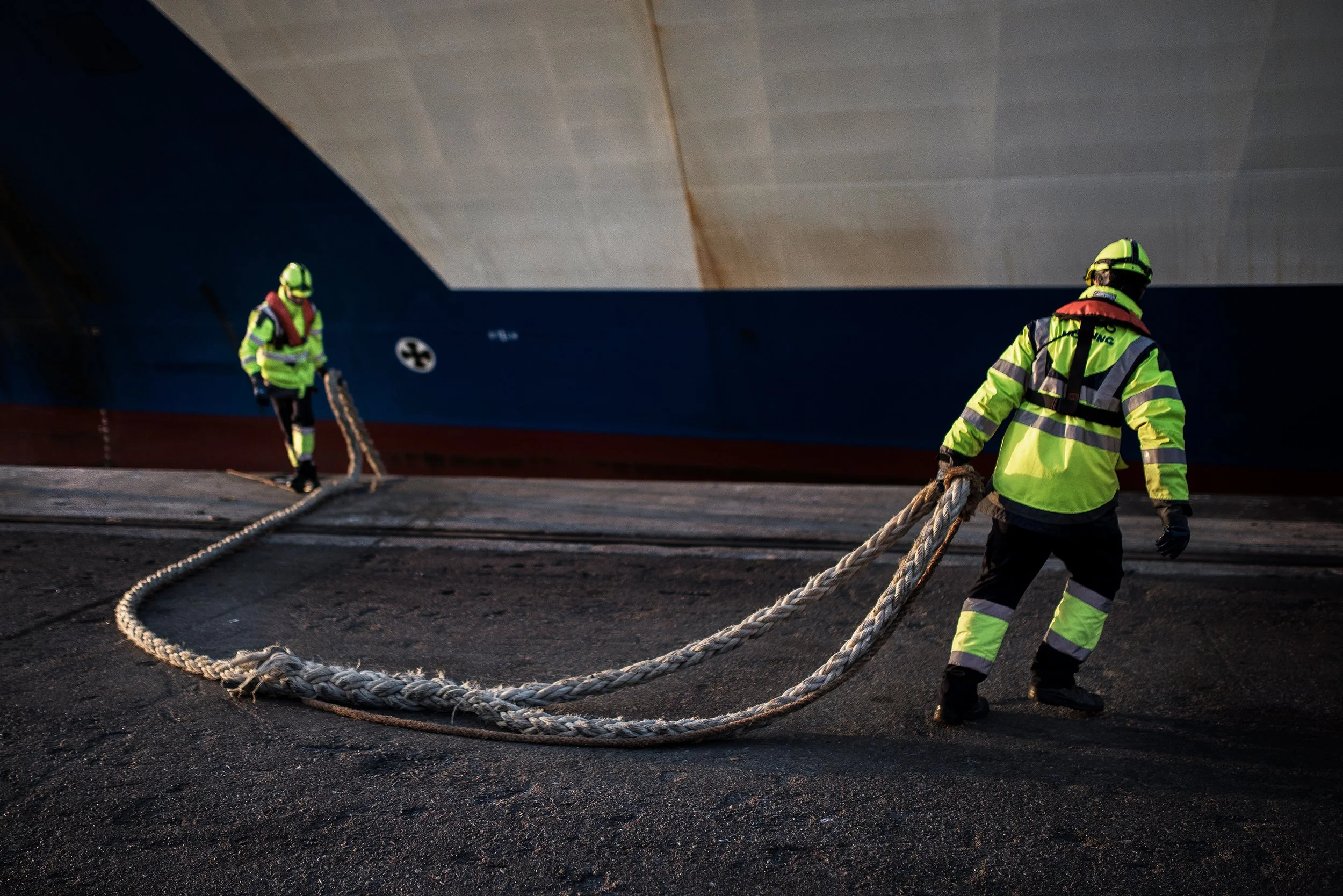 Dos rescatistas con chalecos de alta visibilidad y cascos, sujetando una cuerda gruesa en un muelle de embarcadero.