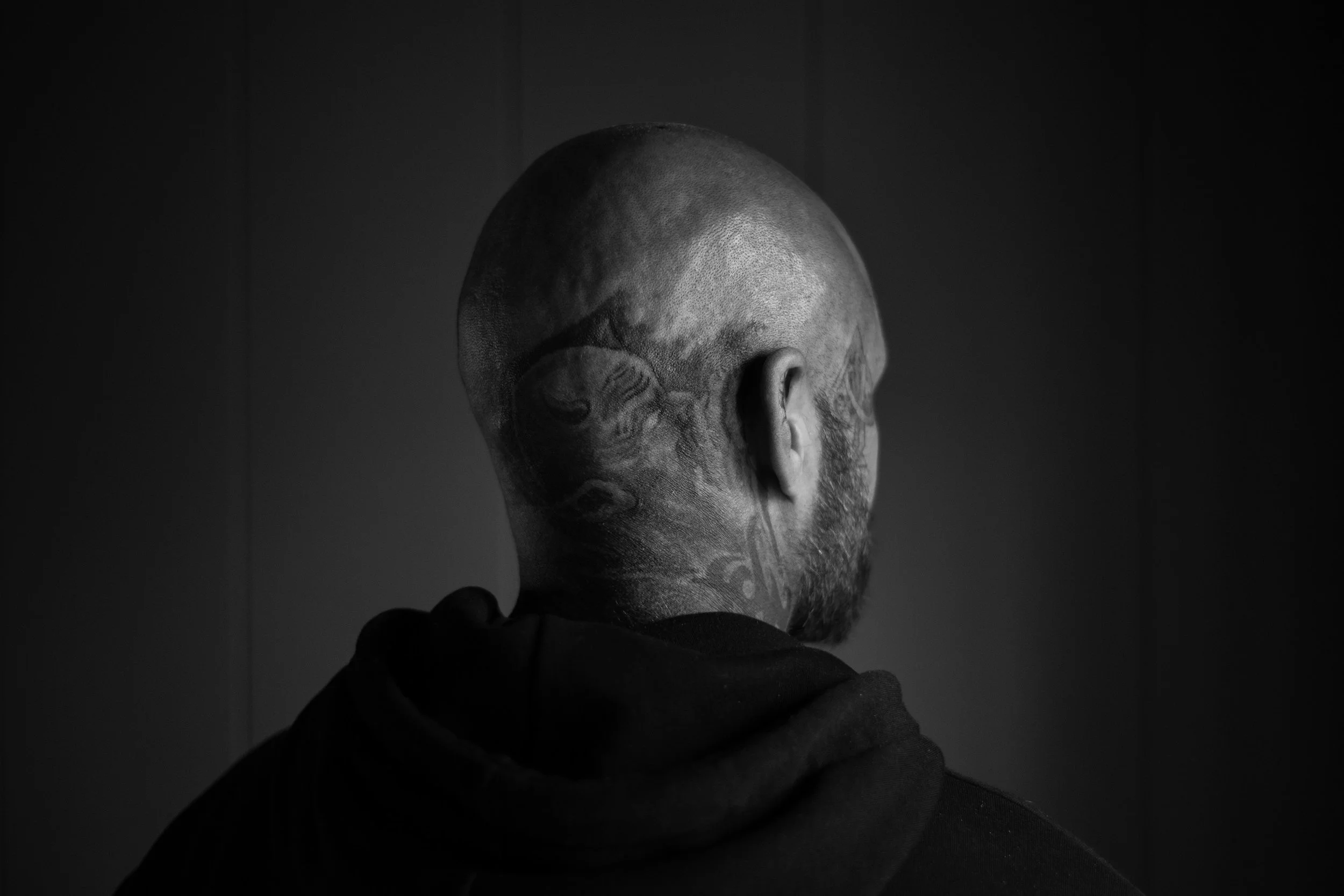 Retrato en blanco y negro de un hombre con cabeza rapada y tatuajes en el cuello, visto desde atrás.