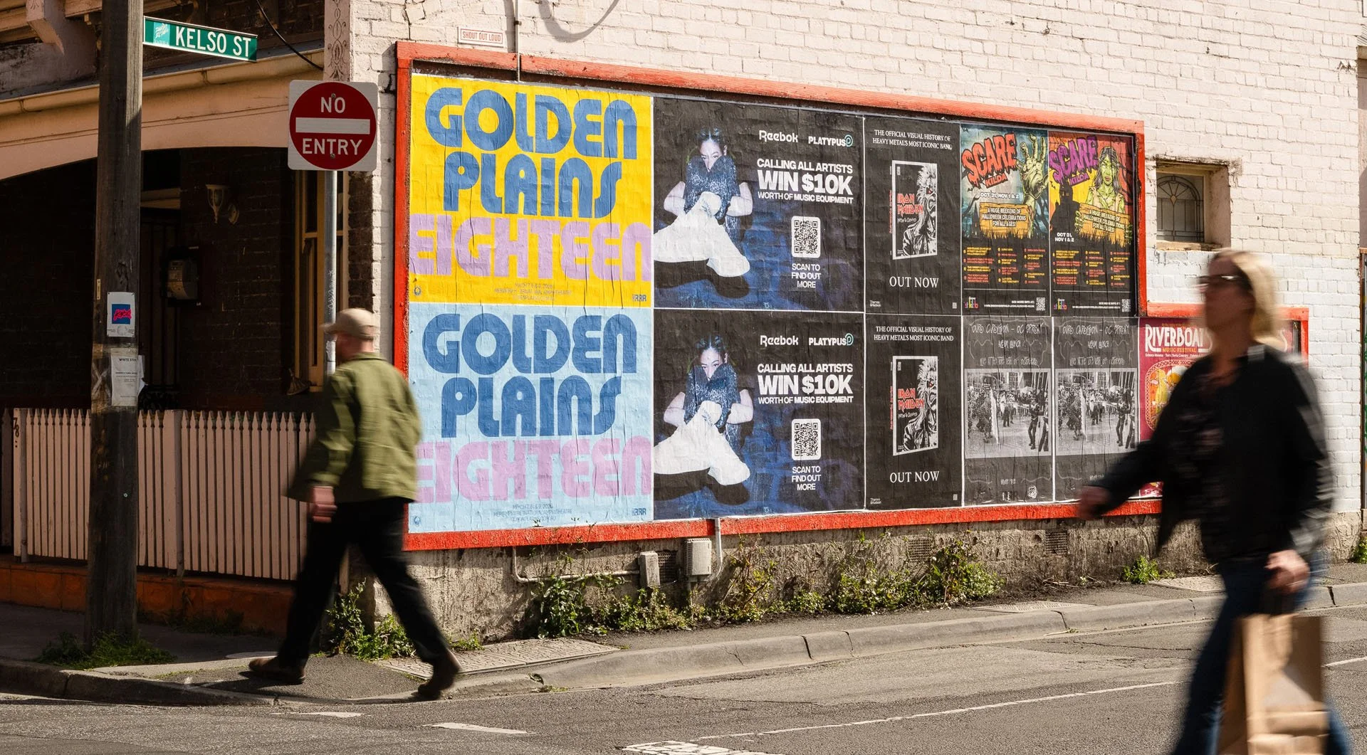 Two pedestrians crossing the street in front of a brick building with large posters on the wall, including advertisements for concerts and events, and a street sign reading 'Kelso St'.