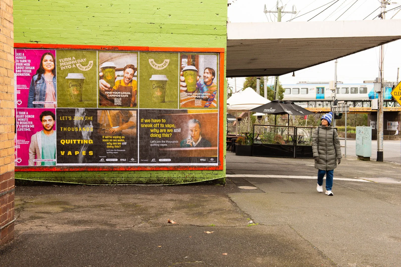 A person in a gray coat with a blue and white beanie walks past a green brick building with three advertisements for Campos coffee and a campaign to quit vaping, on an urban sidewalk near a train station with a train passing in the background.