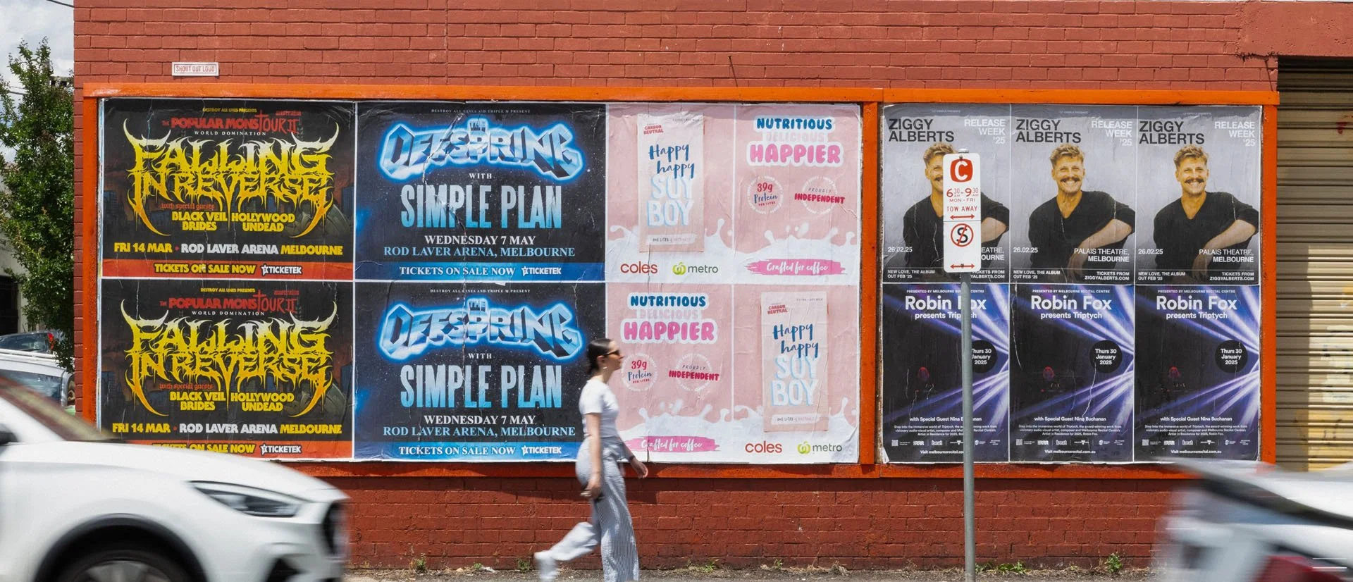 A brick wall with various advertisements and posters, including concerts and event announcements, with a person walking in front and cars passing by.